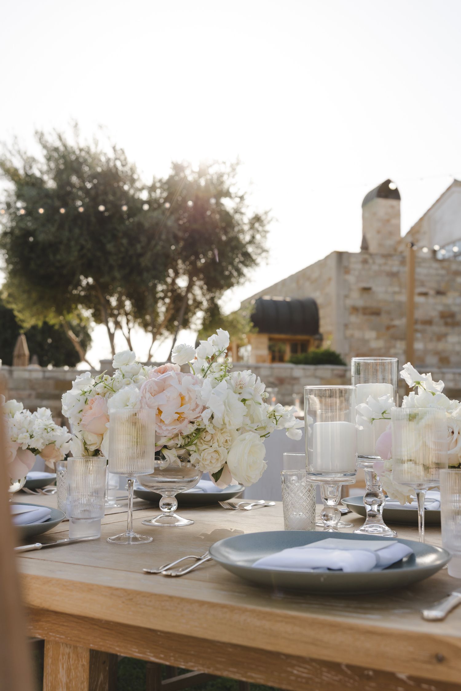 Elegant outdoor table setting with white floral centerpieces, glowing candles and place settings at dusk.