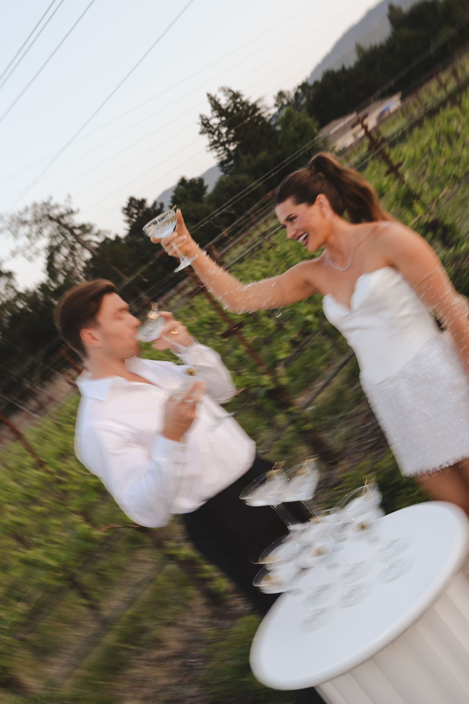 Fun wedding photography showing couple playfully toasting with champagne glasses in an outdoor vineyard setting.