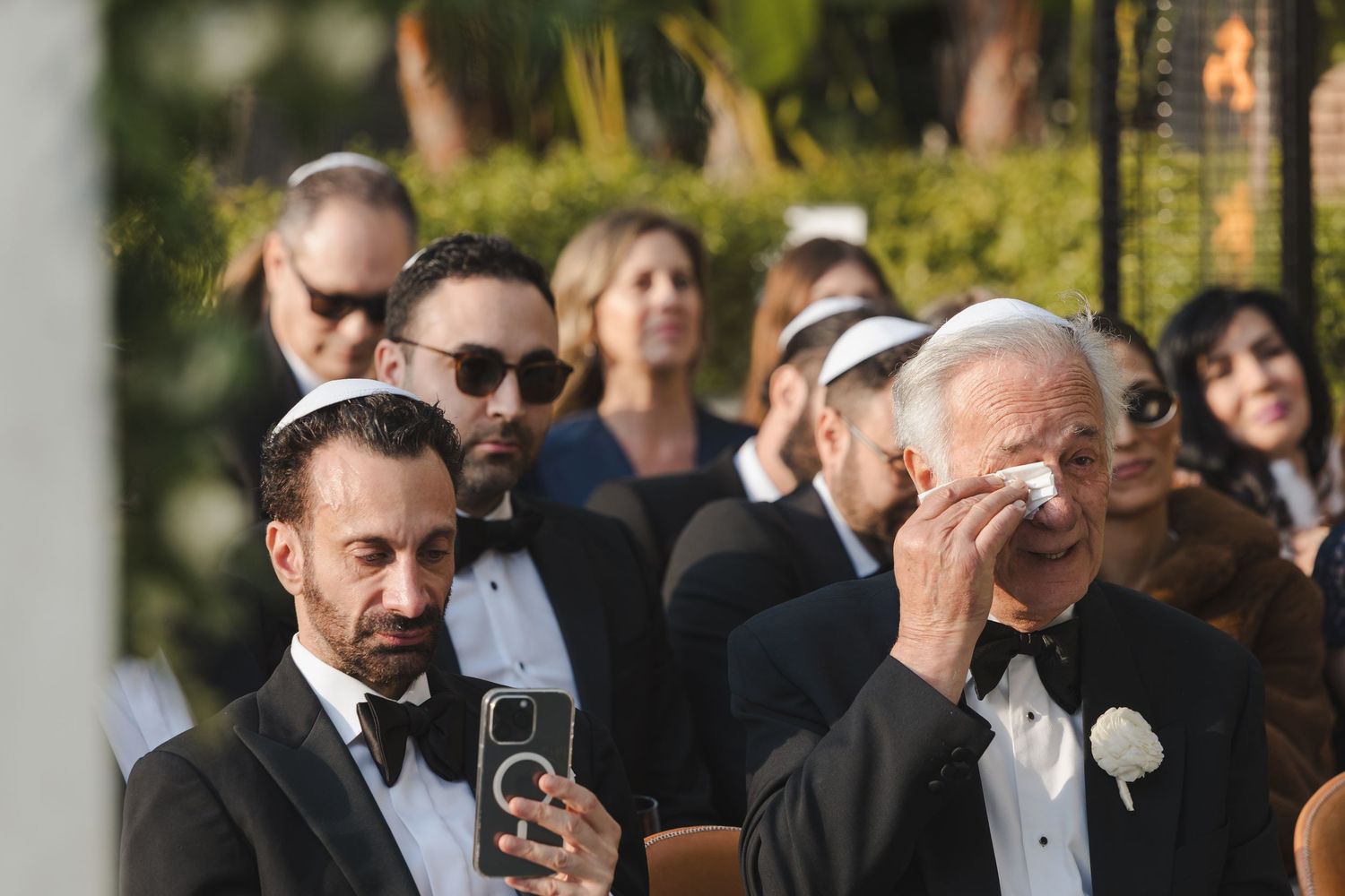Wedding guests in formal black tie attire display emotional reactions during a ceremony outdoors.