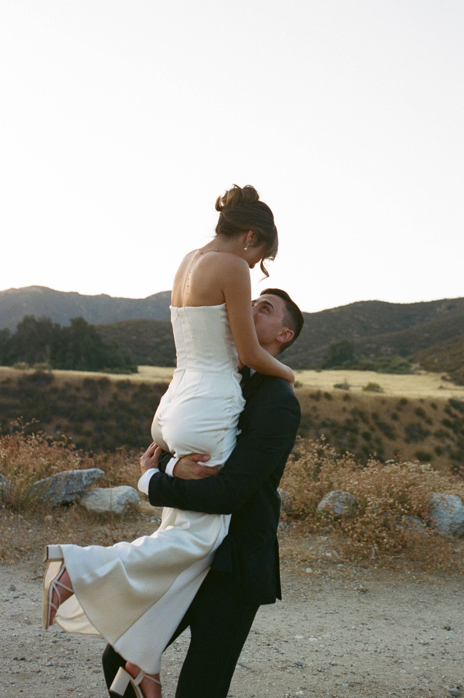 Joyful wedding couple embracing and dancing against a scenic mountain landscape at sunset.
