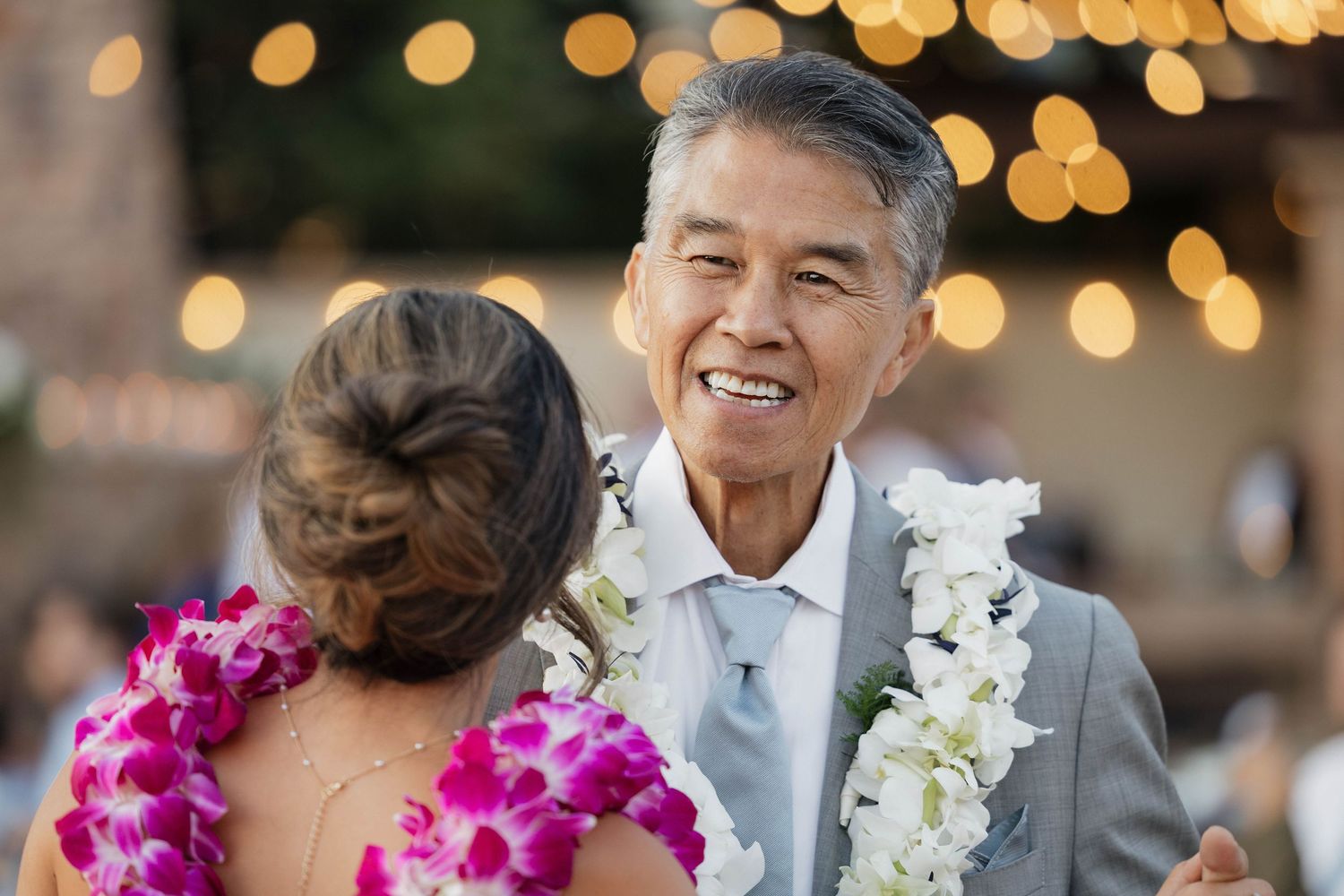 Wedding party celebrates with leis and bouquets under warm string lights.
