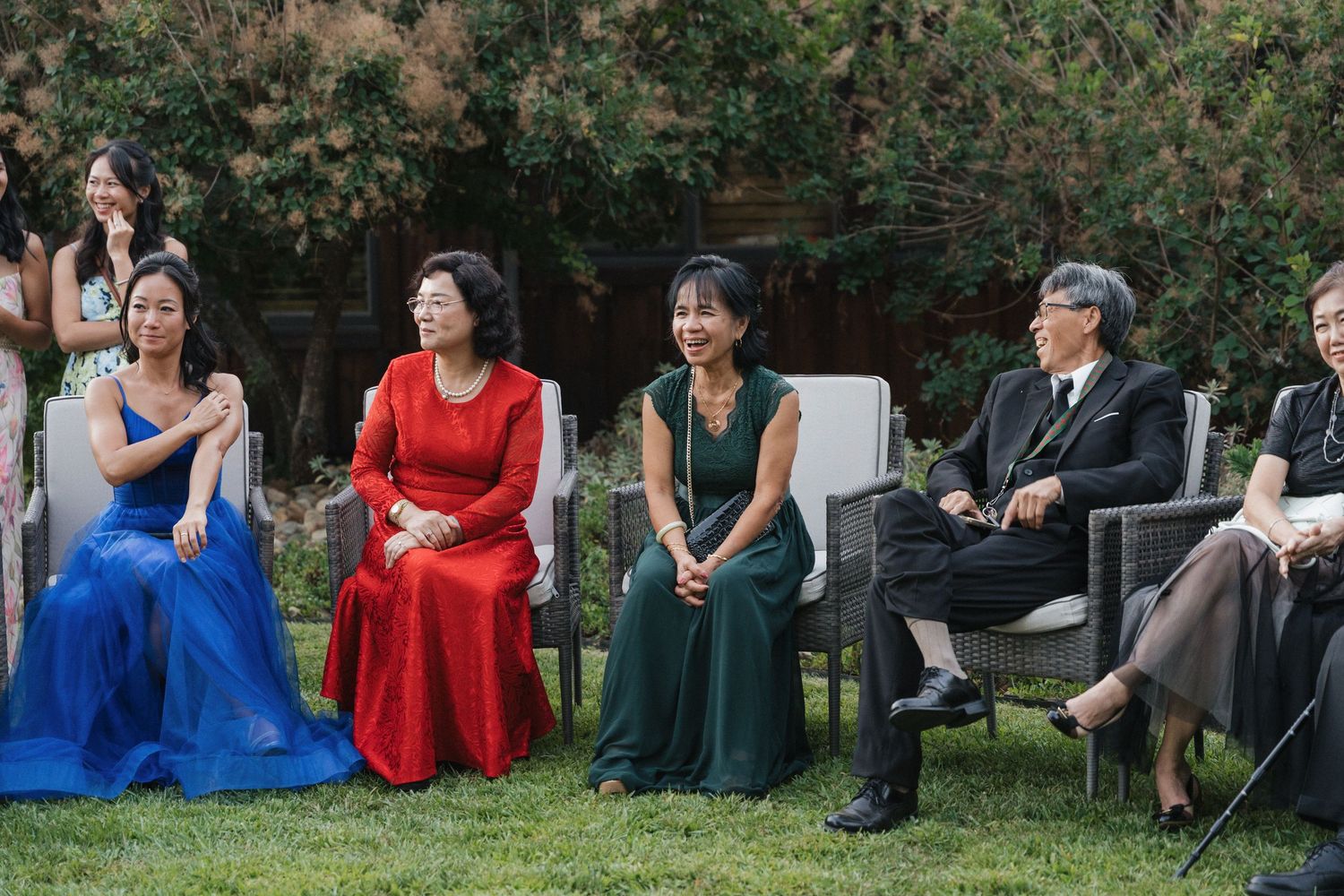 Wedding guests in colorful formal attire sitting on grass during an outdoor celebration.