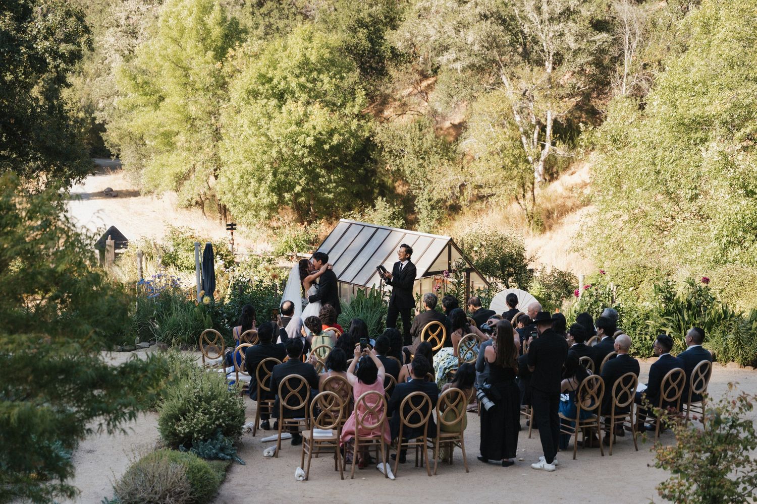 Outdoor wedding ceremony setup with guests seated in rows among trees and natural scenery.