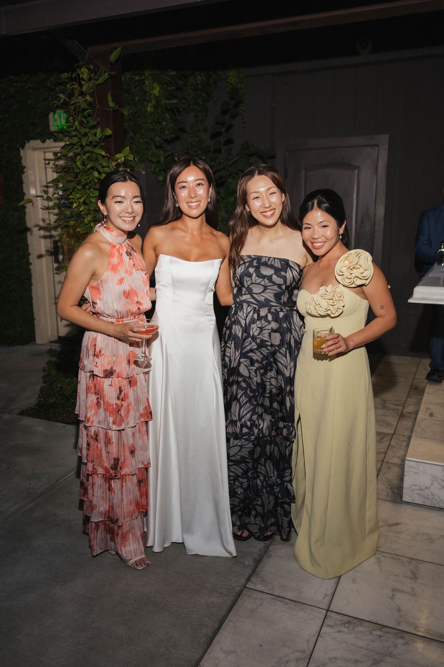 Four women in summer dresses pose together at an outdoor evening event.