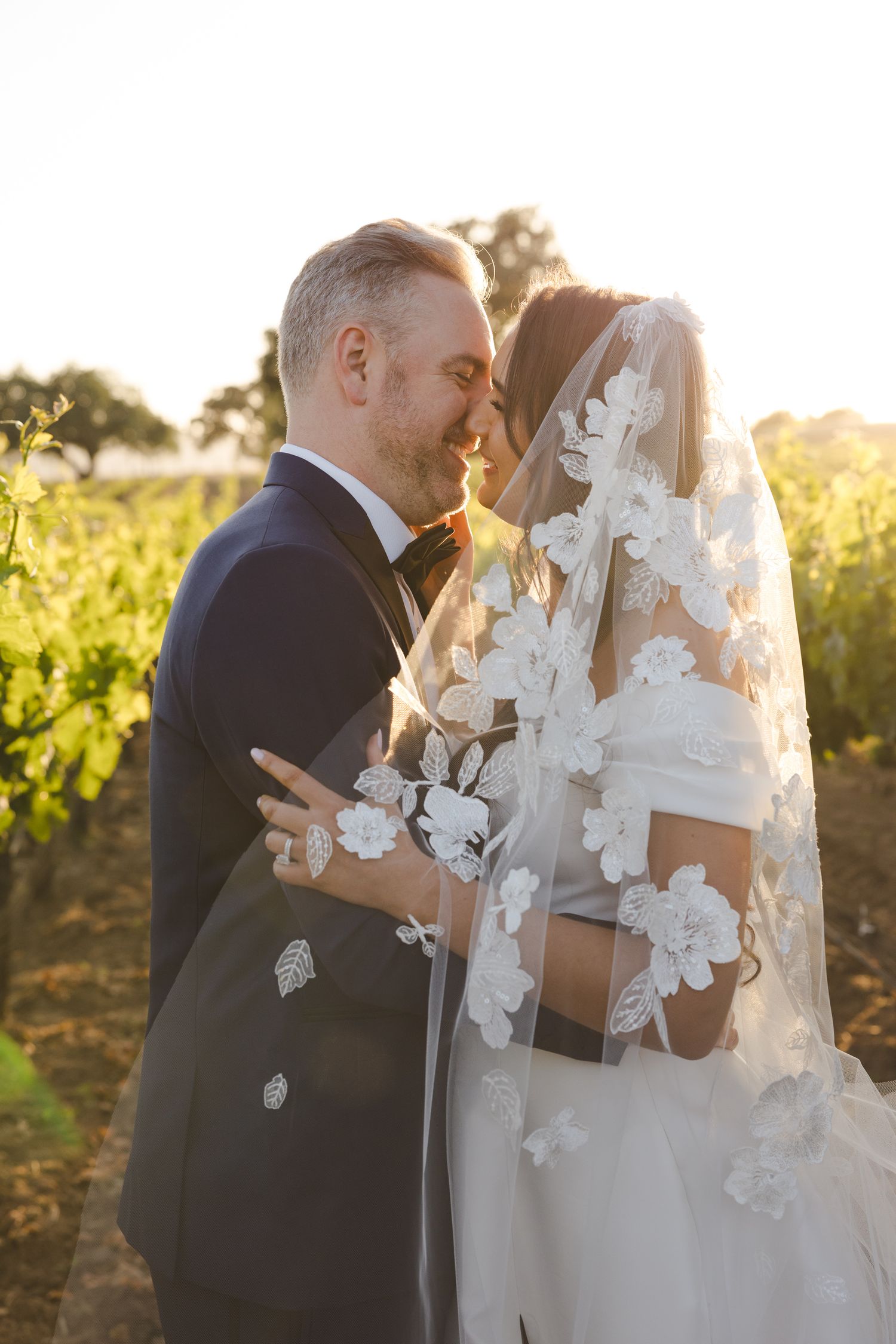 Intimate wedding portrait with delicate butterfly details on veil in golden sunlight.