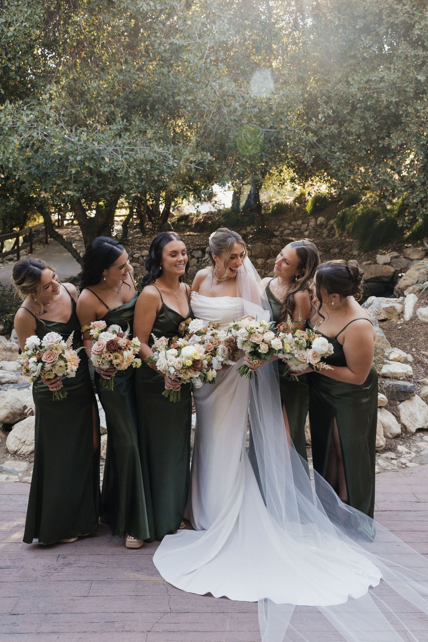 Wedding party in dark green dresses and white gown gather for formal photos in an outdoor garden setting.
