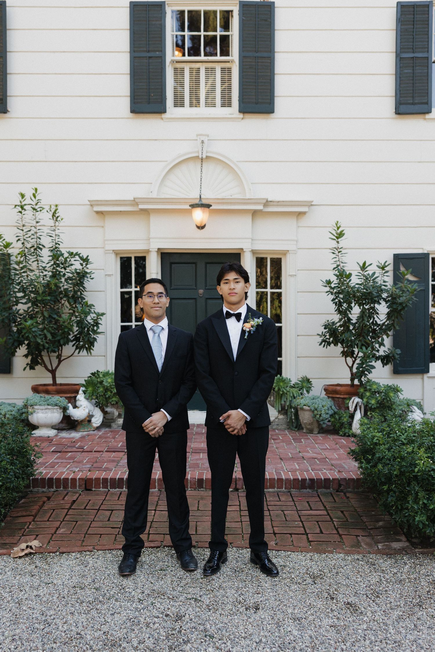Groomsmen in matching black tuxedos pose formally on a gravel path in front of a white mansion.