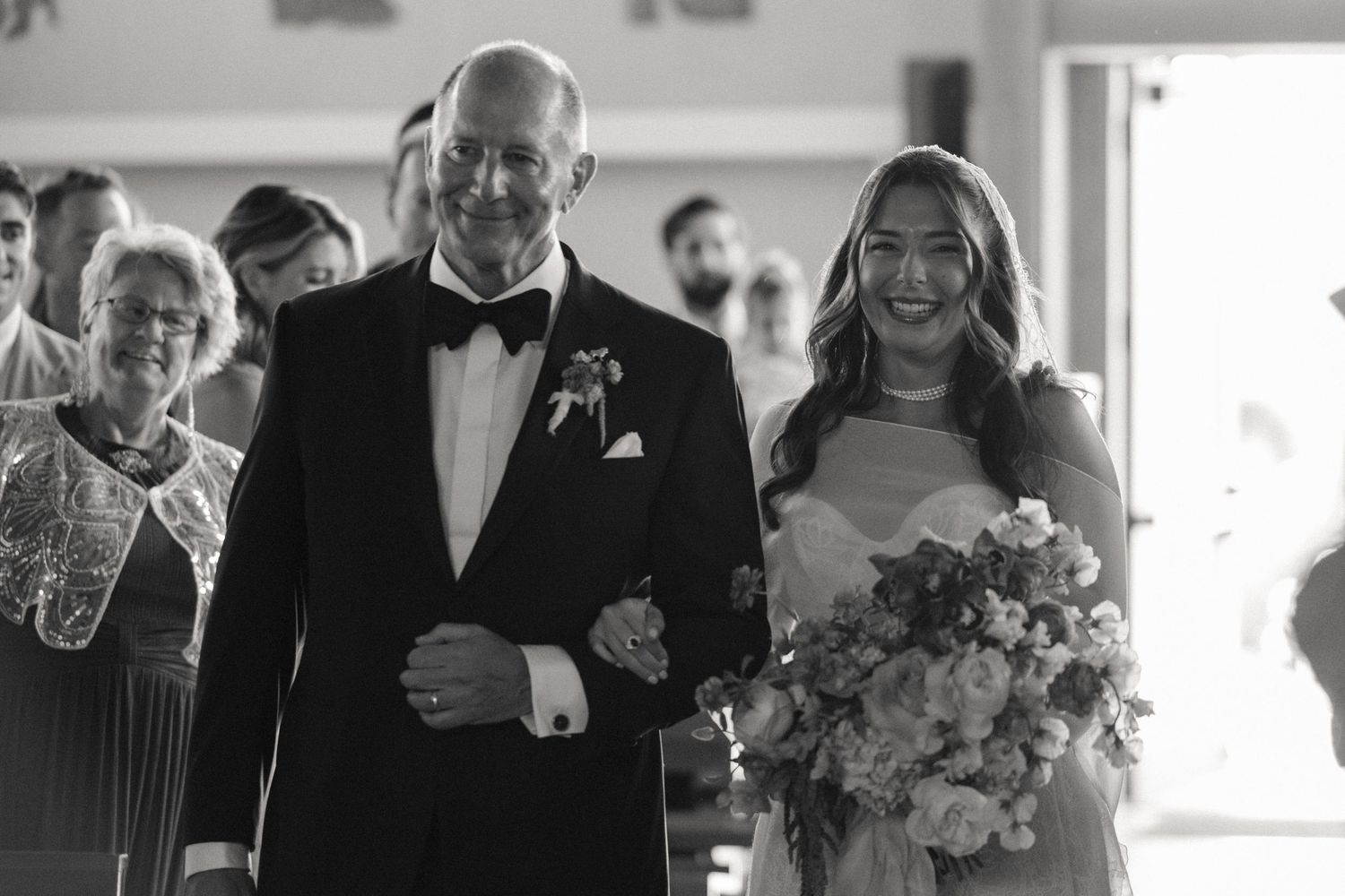 Black and white photo of a bride walking down the aisle with her father at a wedding ceremony.