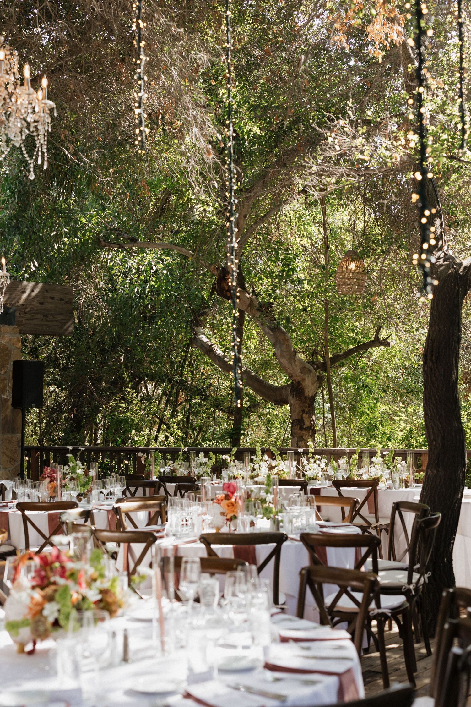 Elegant outdoor wedding reception with string lights, chandeliers, and floral centerpieces under lush trees.