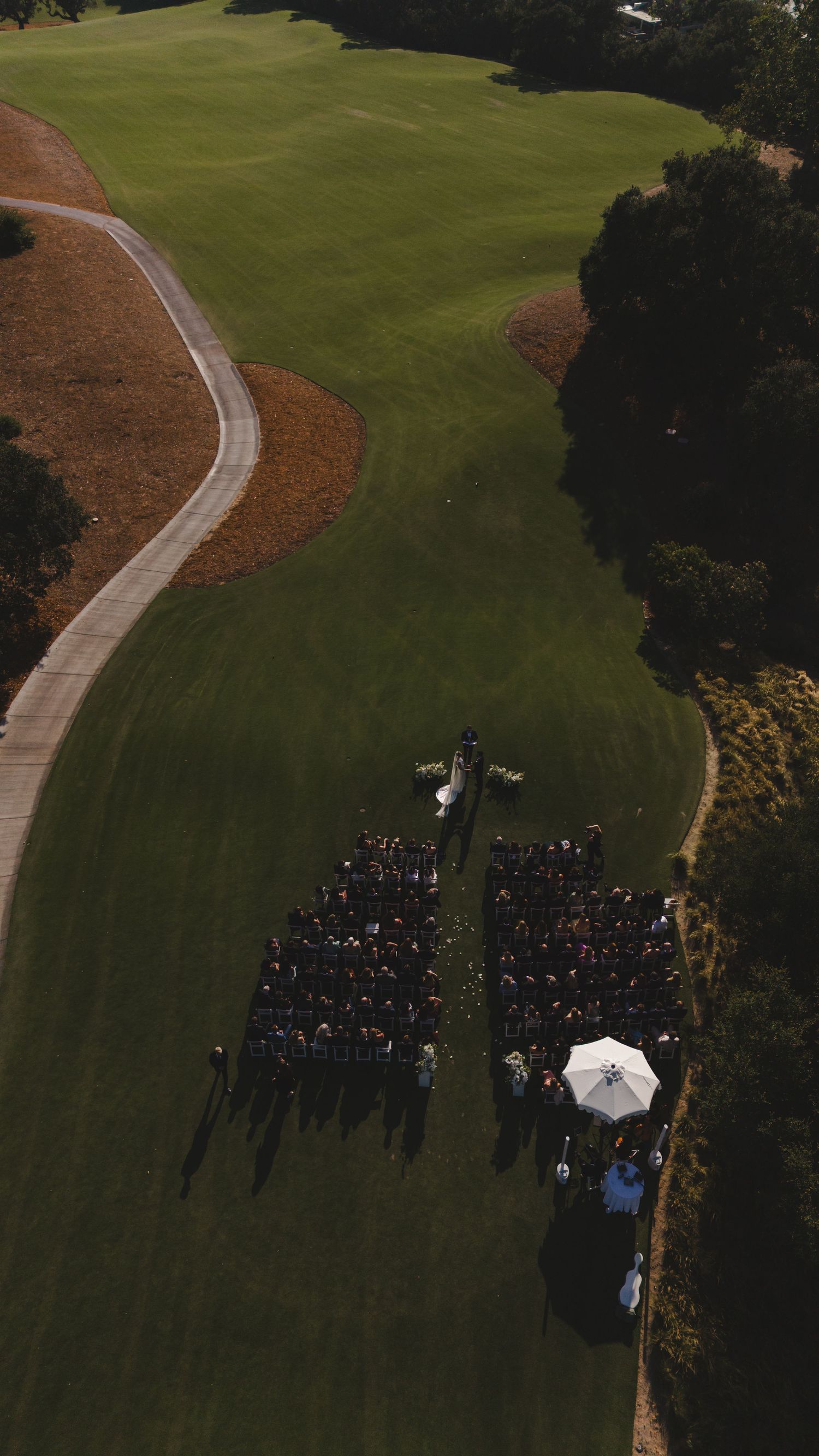Aerial drone view of an outdoor wedding ceremony on a lush green golf course with seated guests and a white umbrella.