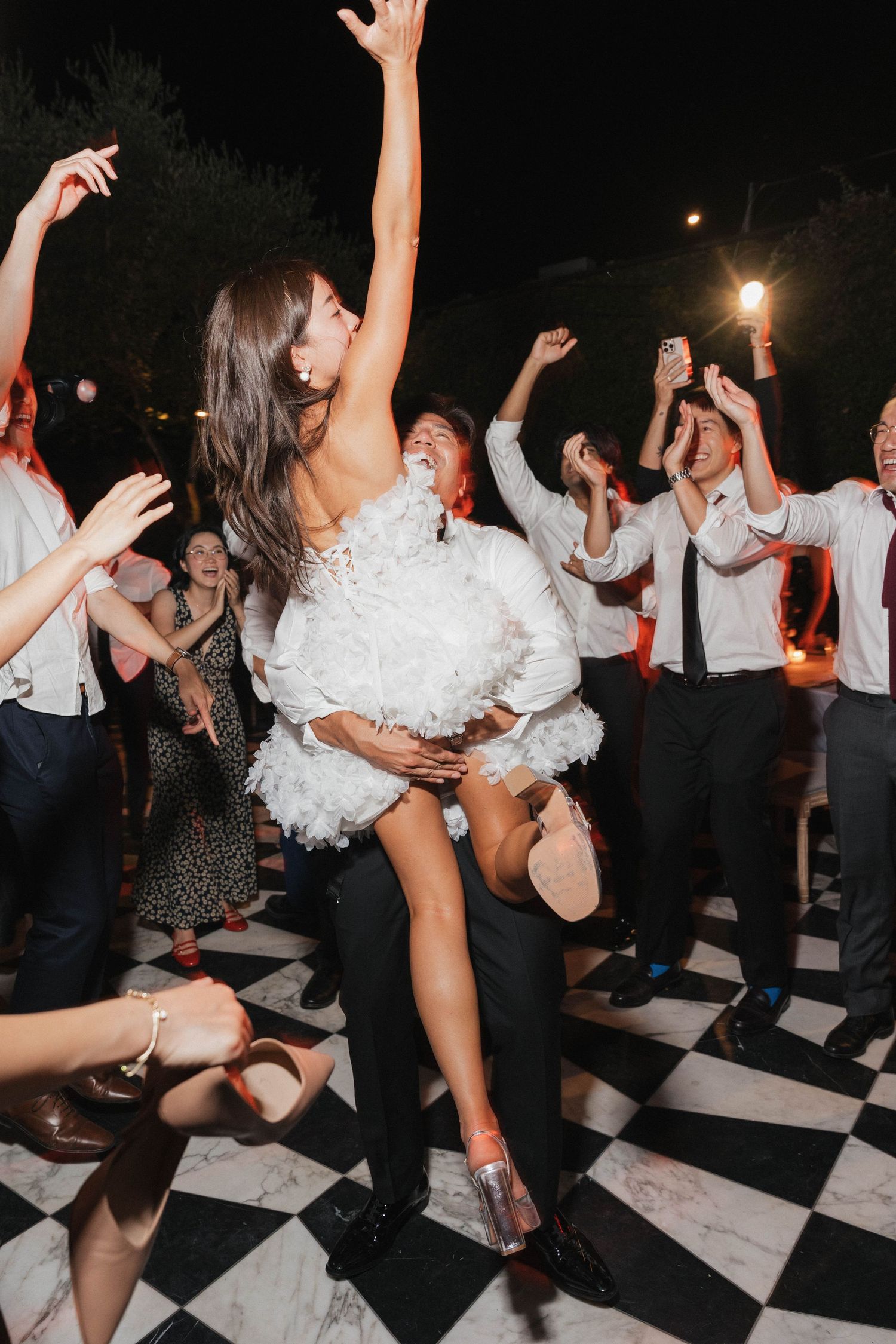 Wedding party guests dance enthusiastically on a black and white checkered dance floor at a nighttime reception celebration.