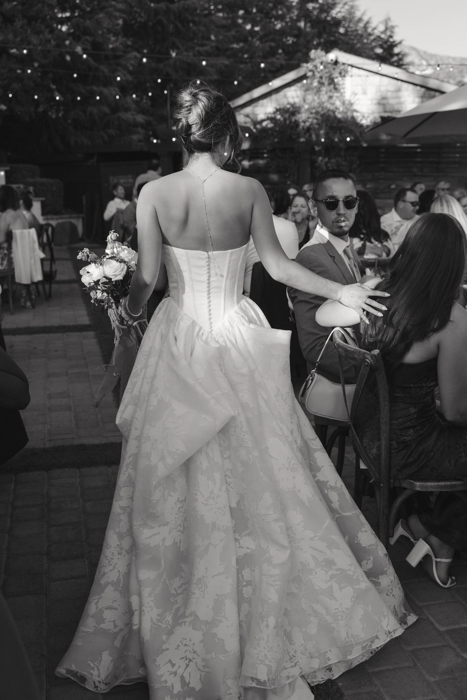 Black and white wedding photo showing the back of a bride in a lace corset gown greeting guests at an outdoor reception.