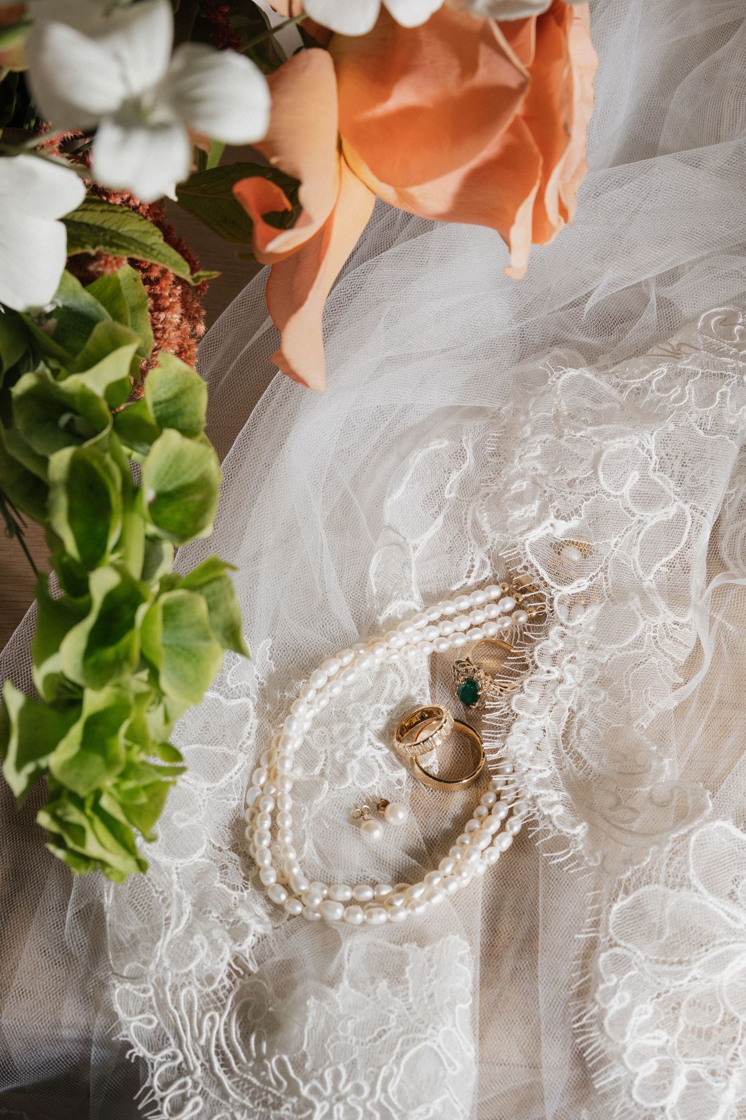Bridal accessories flat lay featuring pearl necklace, gold rings with emerald stone on white lace wedding dress with flowers.