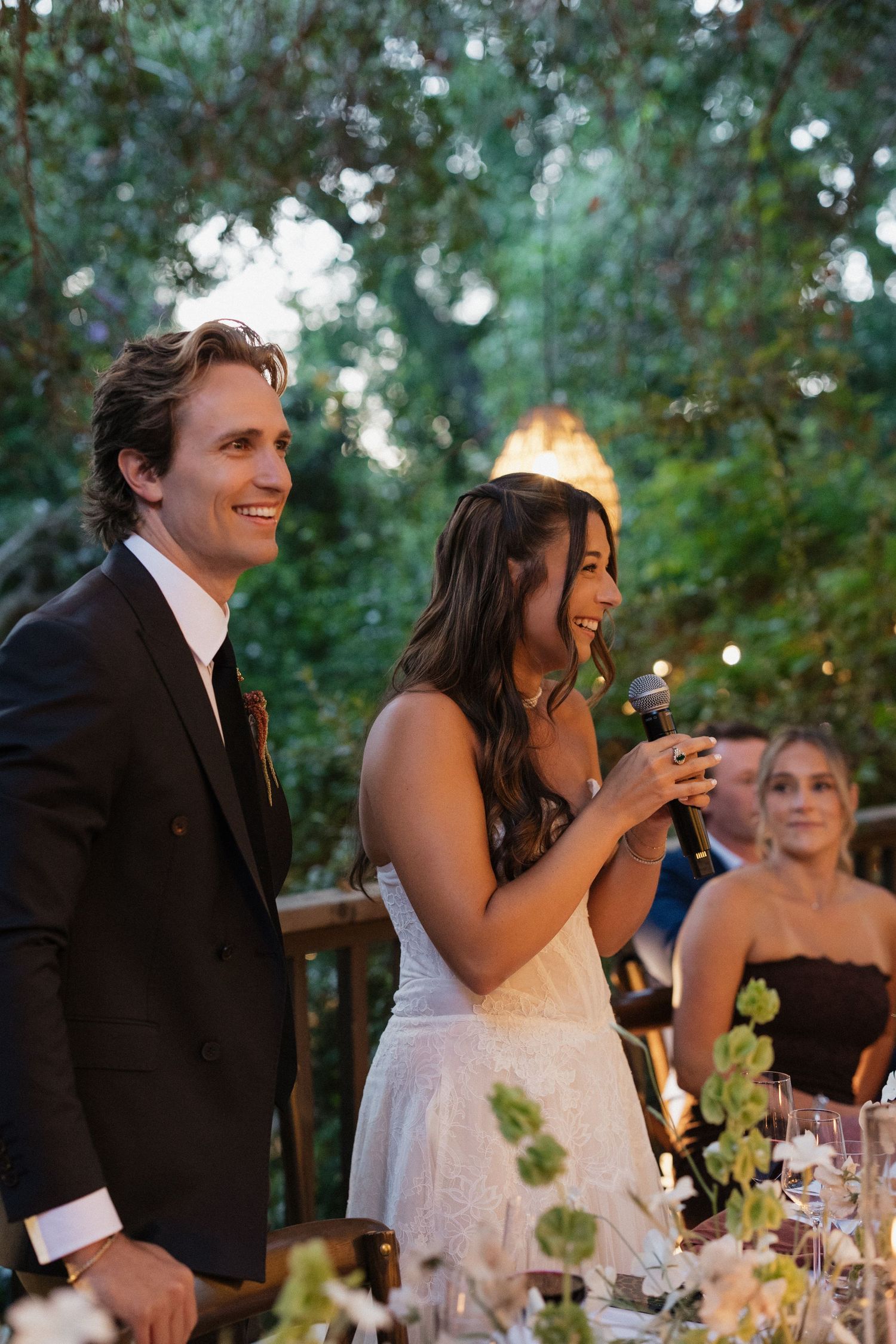 Smiling bride speaks into microphone beside groom at candlelit outdoor wedding reception dinner.