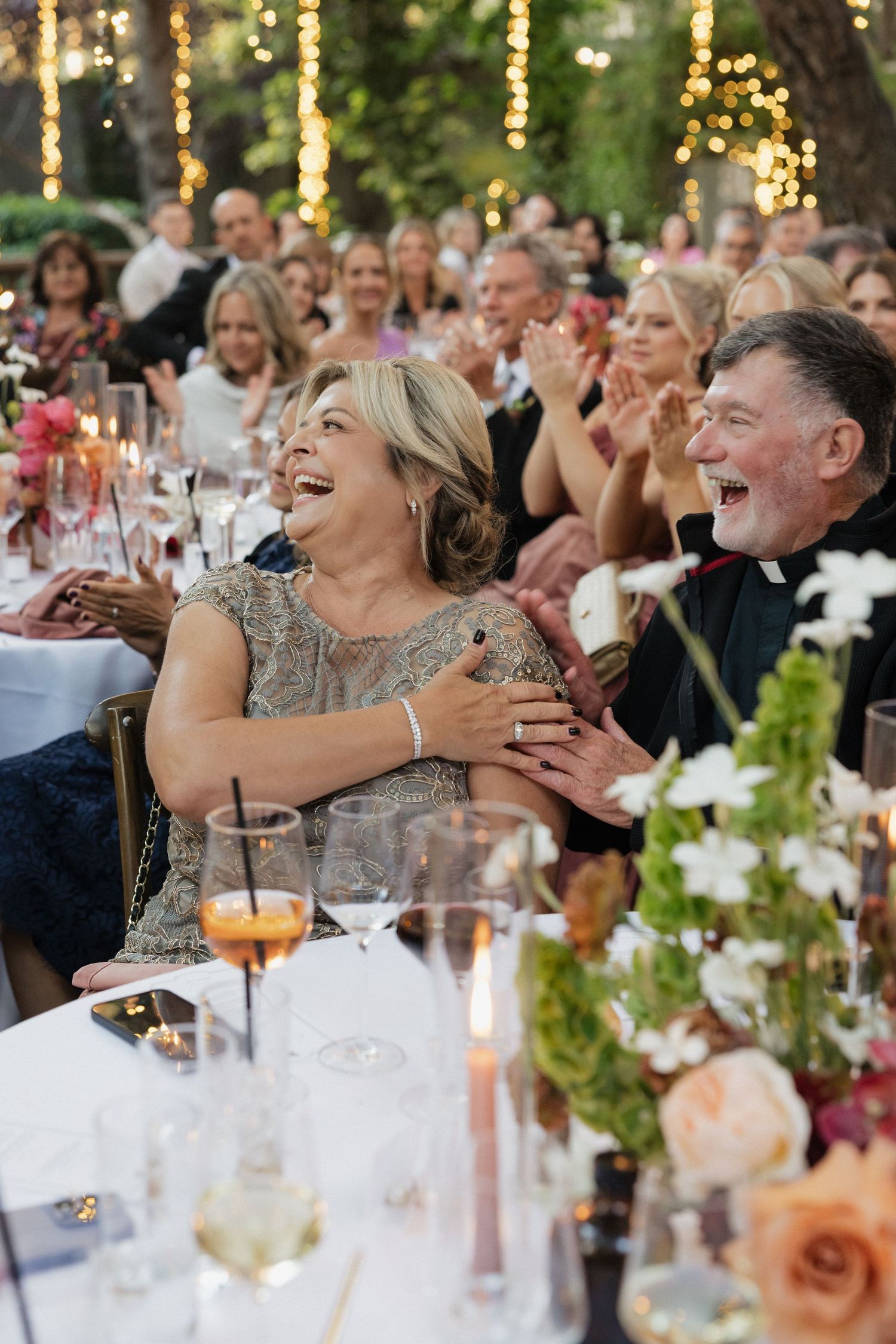 Wedding guests laugh and celebrate at elegantly decorated outdoor reception dinner tables with candles and floral arrangements.