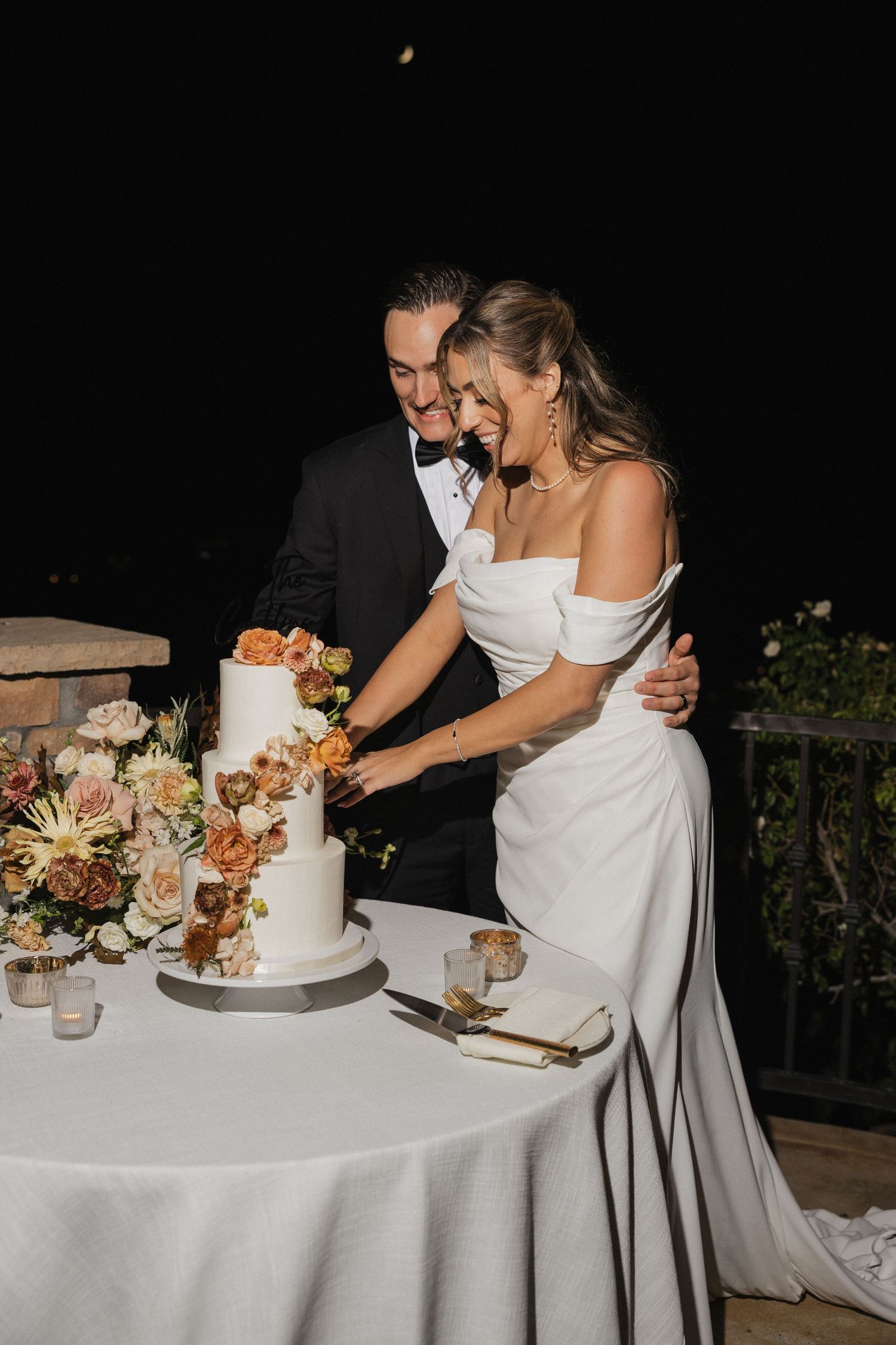 Wedding couple cuts elegant white cake decorated with fresh flowers at reception.