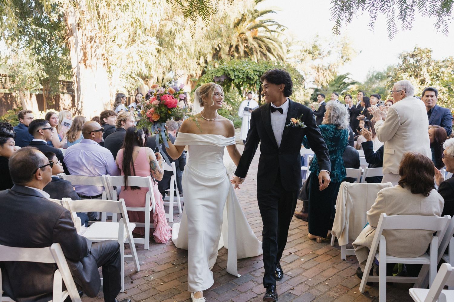 A joyful couple walks back down the aisle together after their outdoor garden wedding ceremony surrounded by guests.
