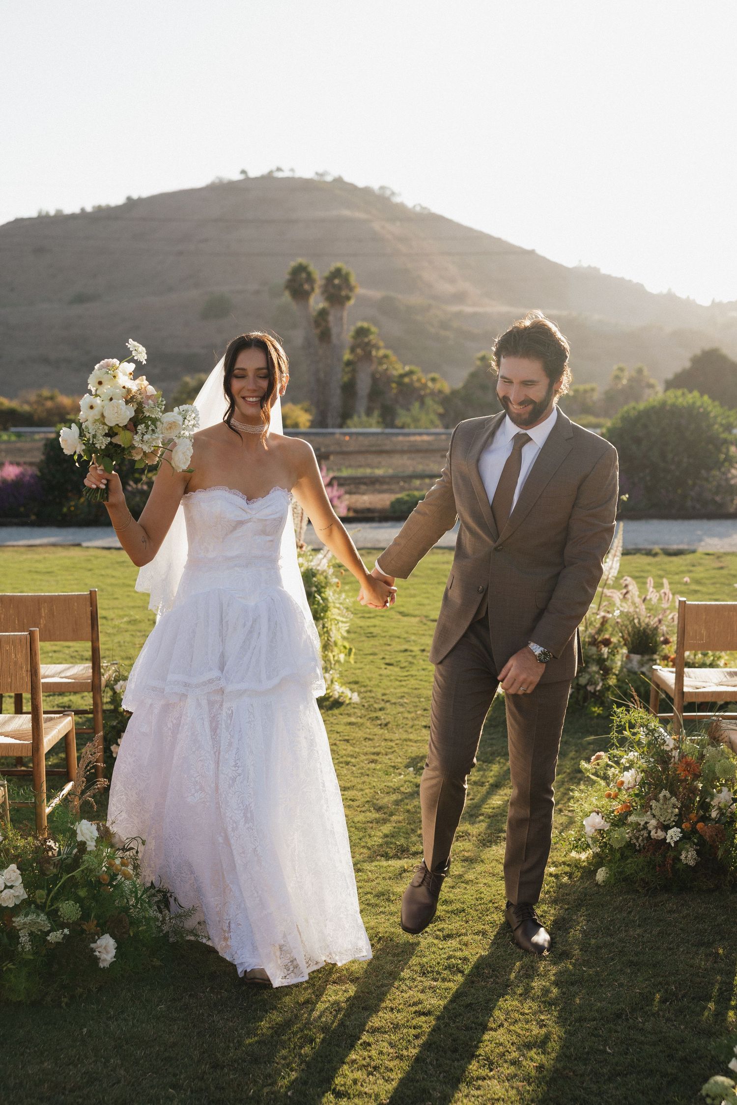 A couple celebrates their wedding outdoors on a grassy lawn with wooden chairs and a scenic mountain backdrop at sunset.