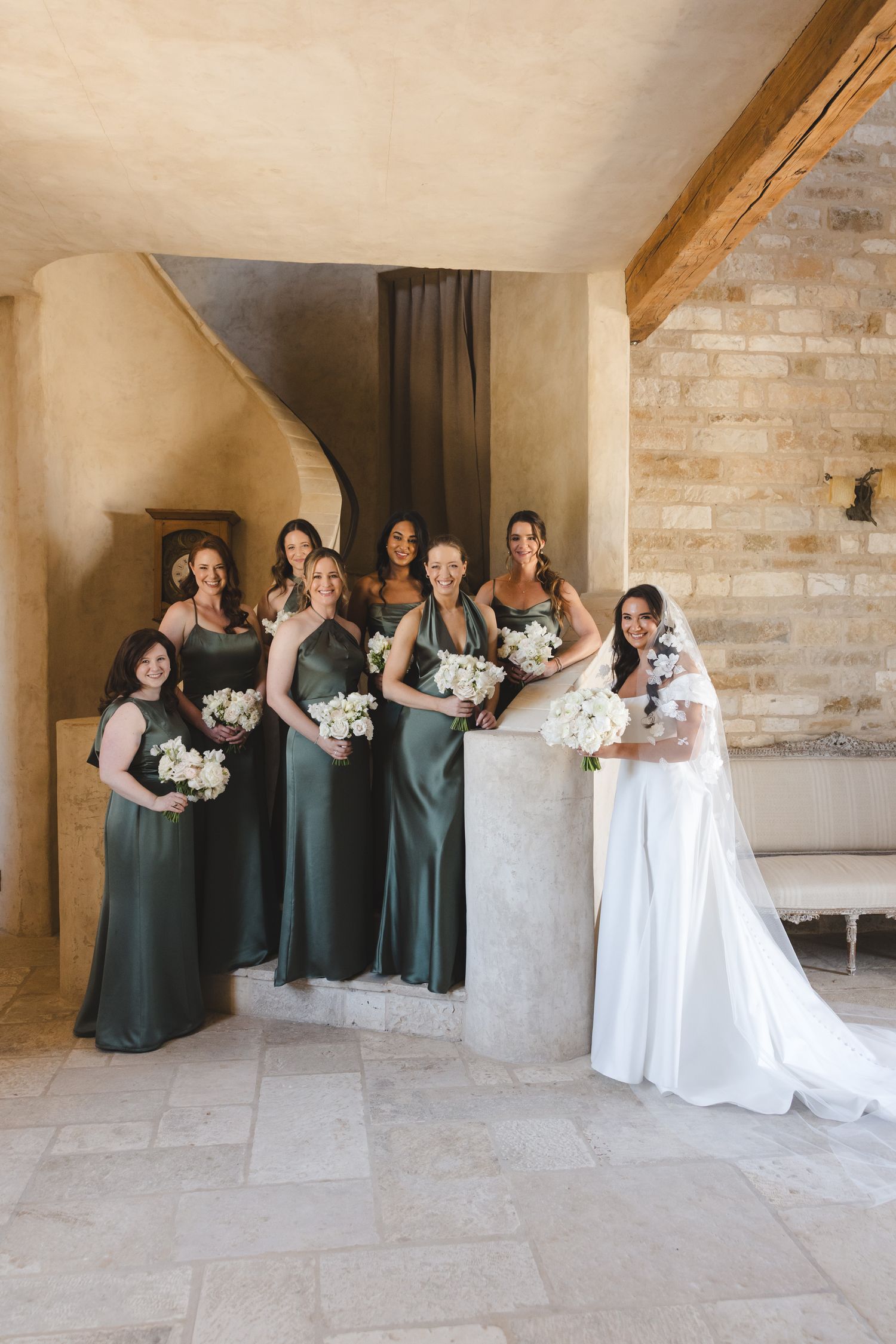 Bridal party wearing sage green dresses poses along a stone wall for wedding photos.