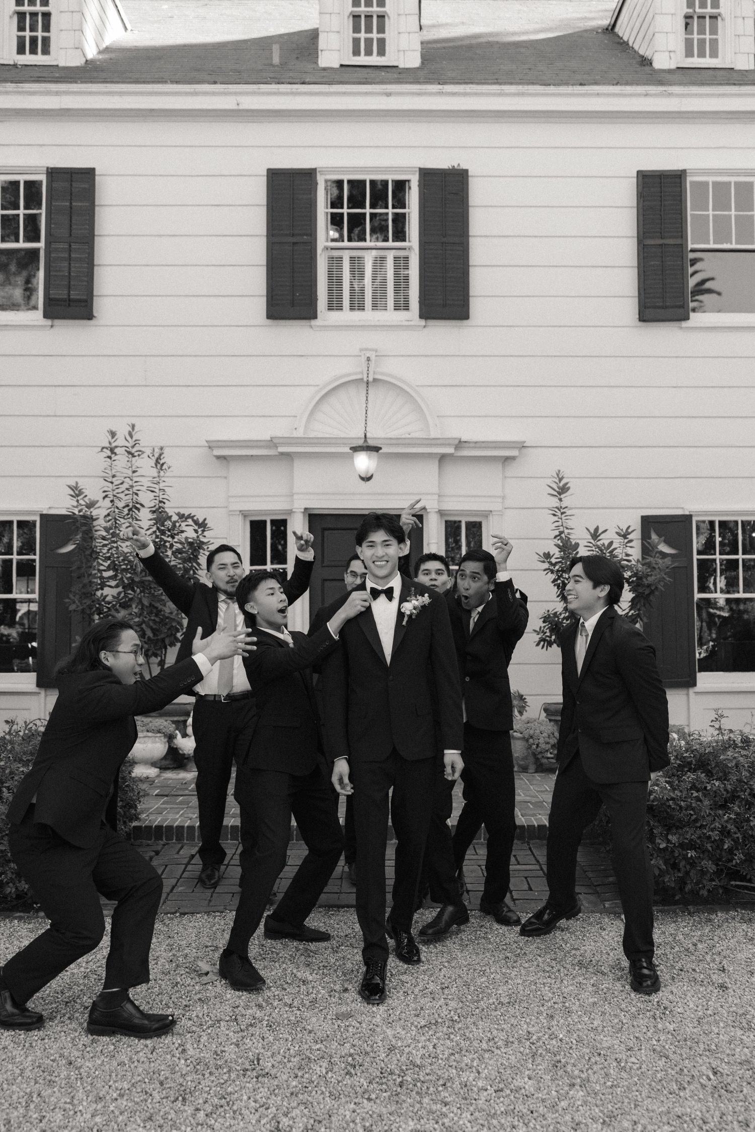 Groomsmen in formal attire pose playfully in front of a white colonial style building.