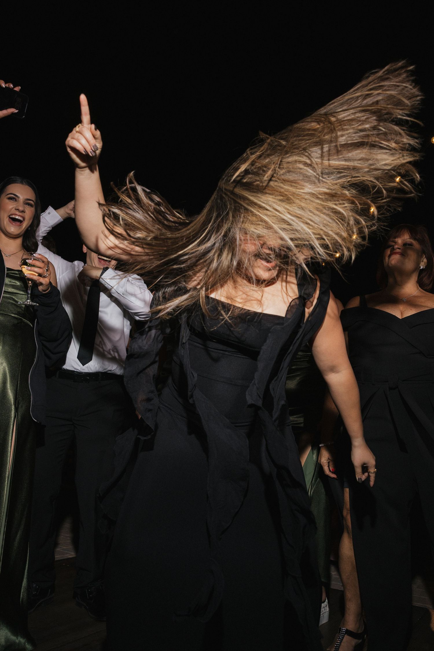 Wedding party members dance energetically with hair flying during nighttime reception celebrations.