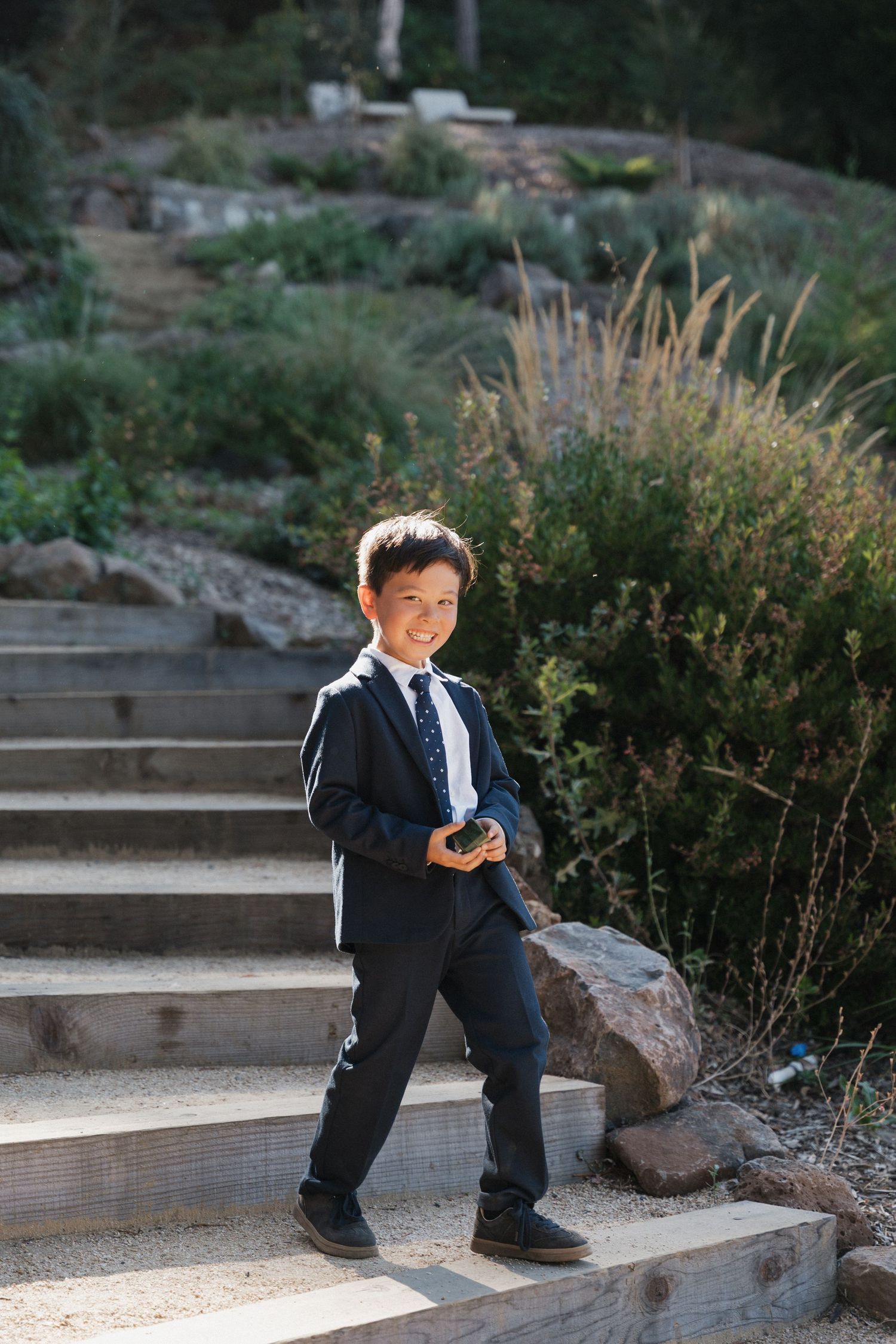 A young boy in a formal suit stands on outdoor stone steps surrounded by greenery.