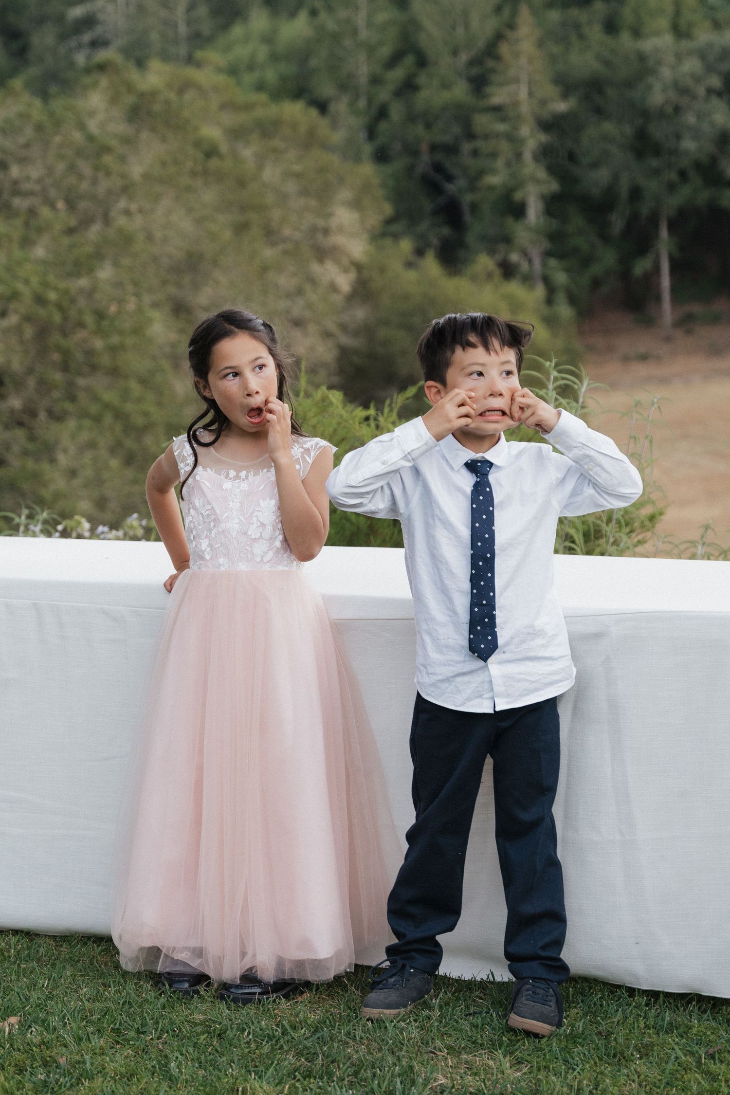 Young children in formal attire playing and posing against white wall at wedding.
