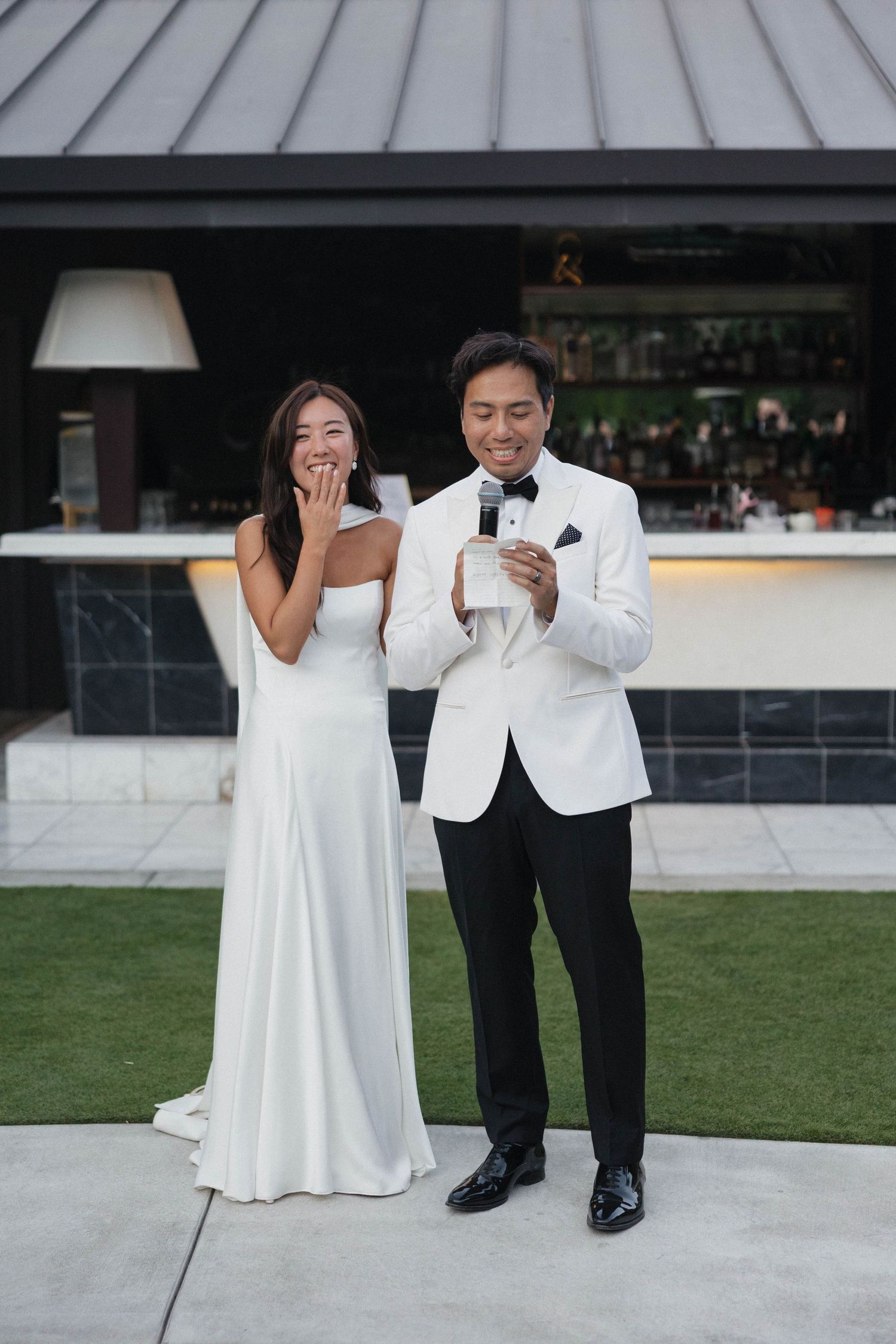 Couple in matching white formal attire poses playfully on outdoor patio.