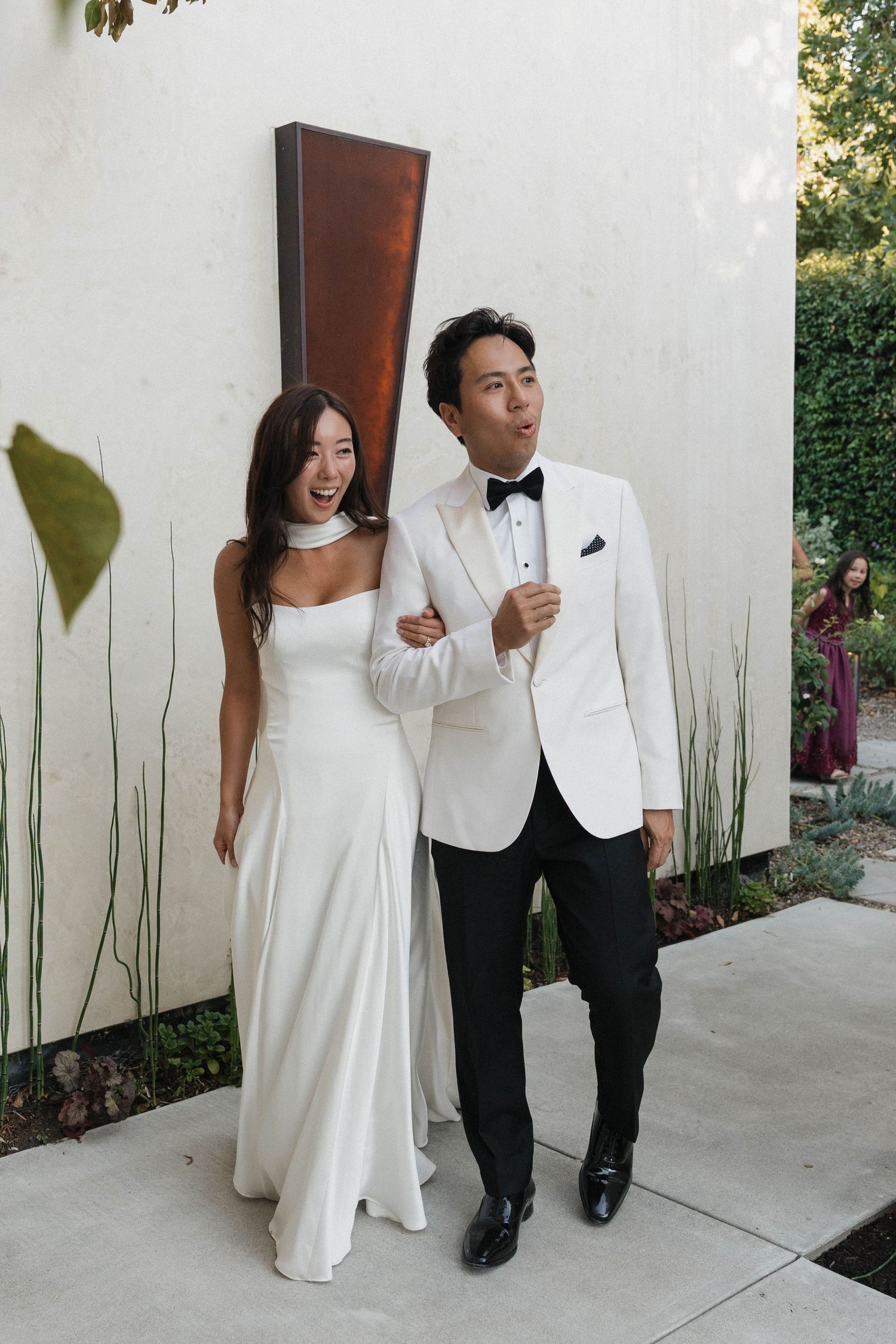 Couple in white formal attire poses playfully against white wall with garden backdrop.
