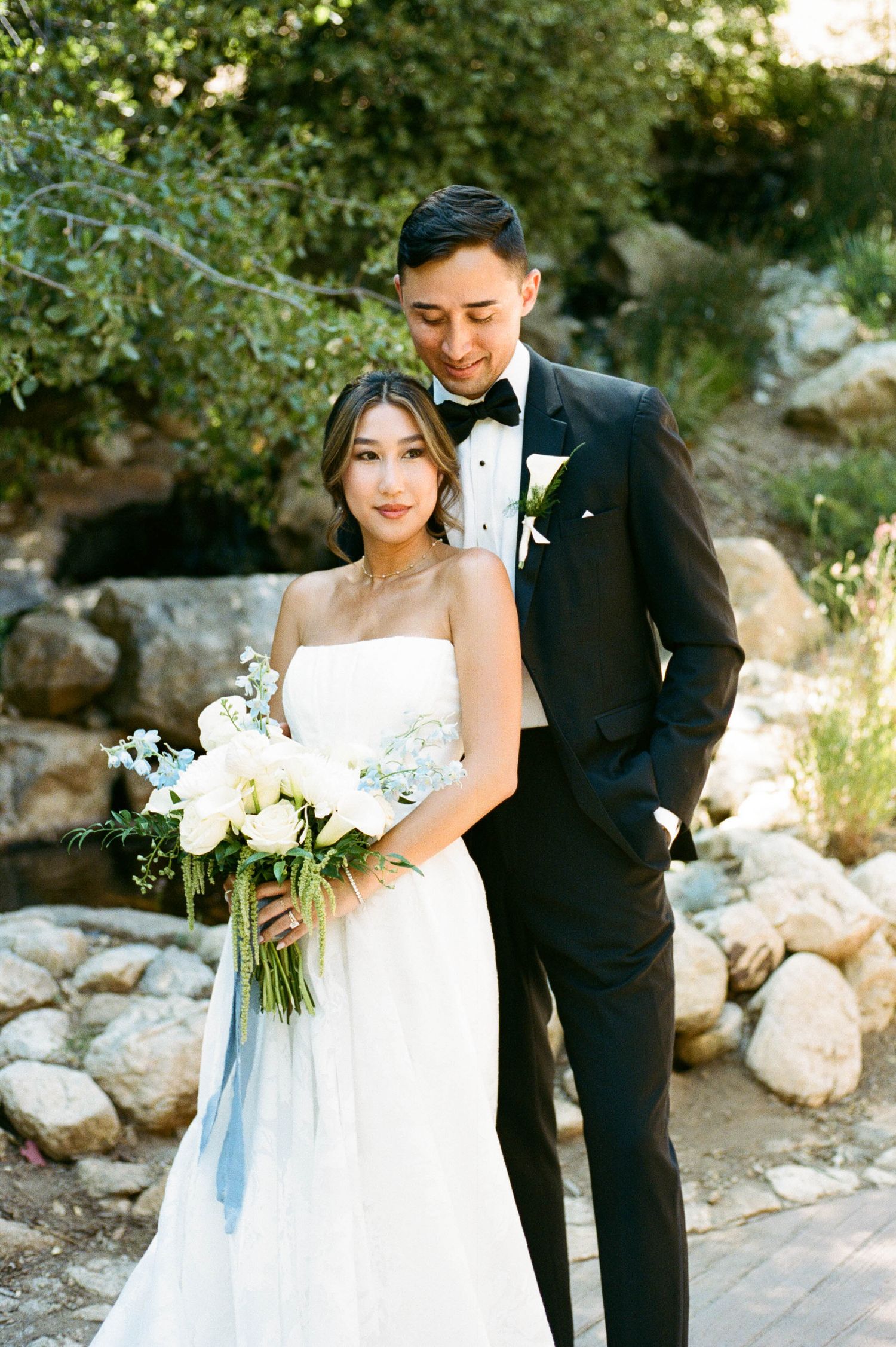 A bride in a white gown and groom in black tuxedo share romantic moments during an outdoor photoshoot.