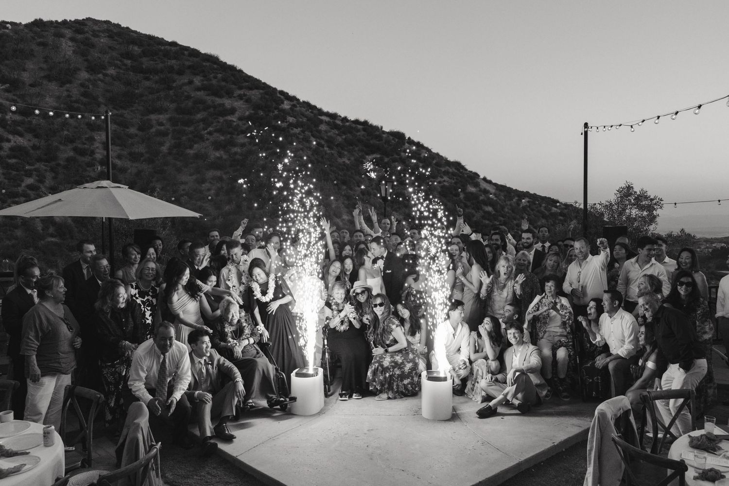 Large wedding reception crowd dancing under string lights at an outdoor mountain venue at dusk.