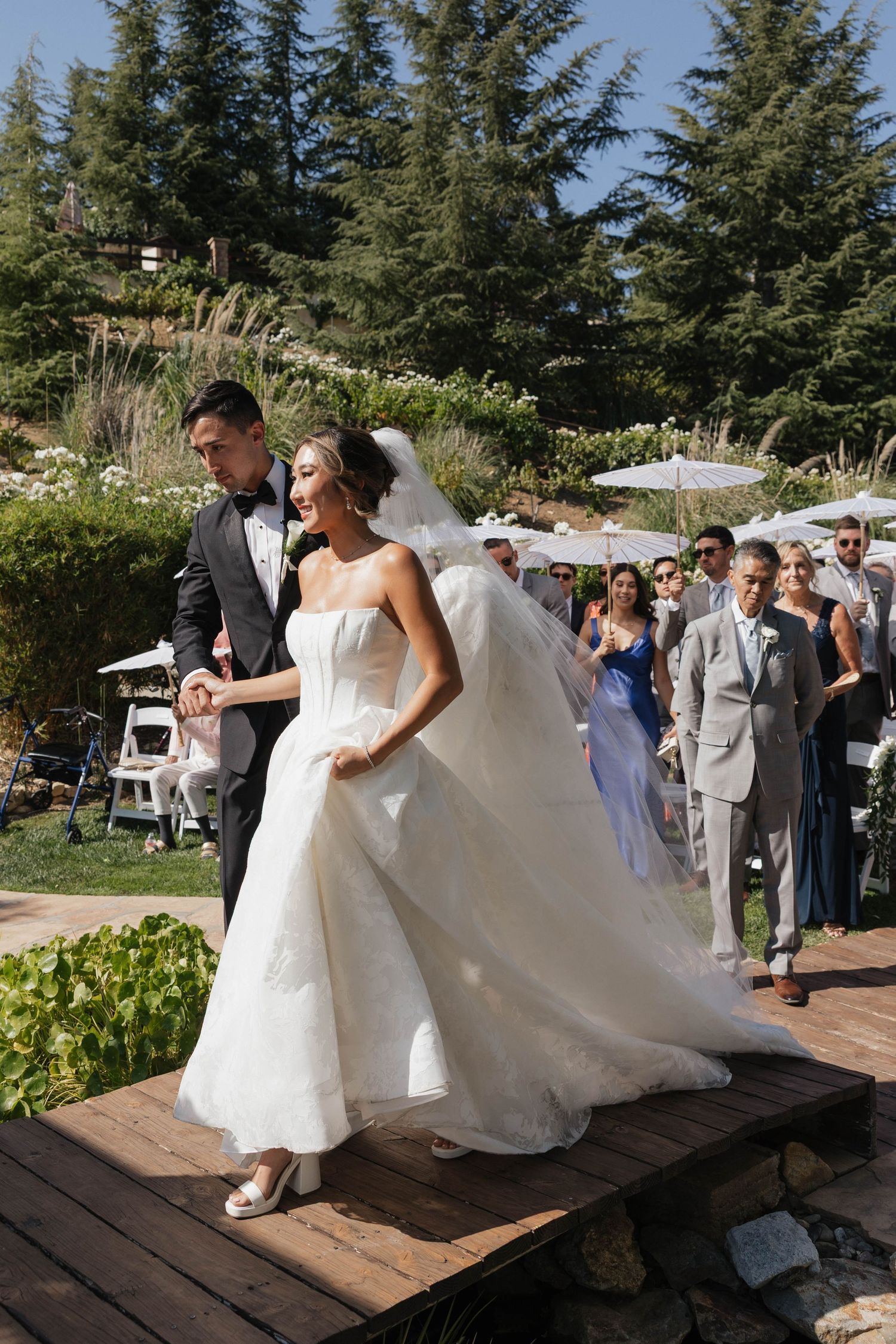 Couple performs first dance on outdoor wooden platform surrounded by wedding guests.