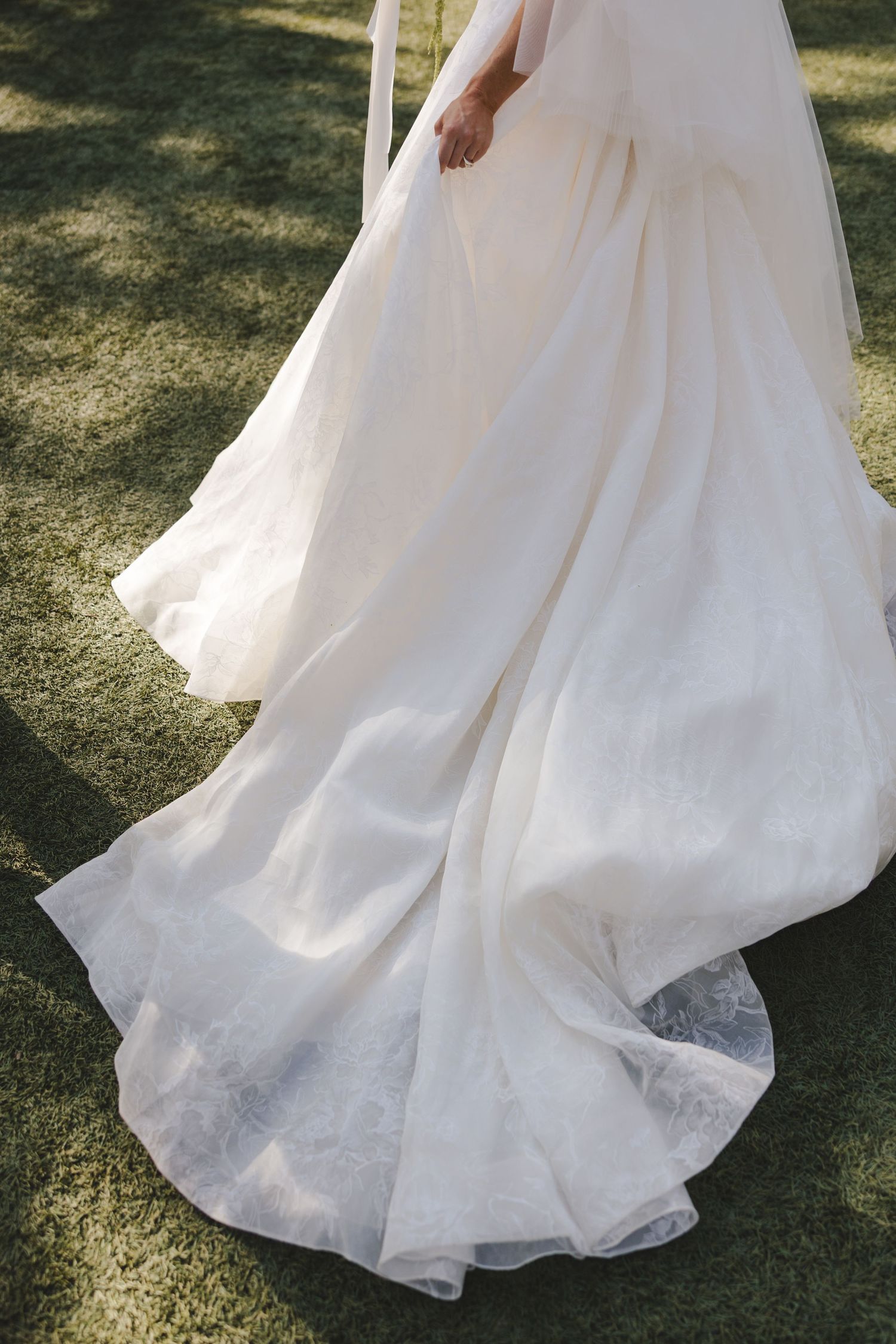 A flowing white wedding dress with delicate tulle fabric trails elegantly across sunlit grass.
