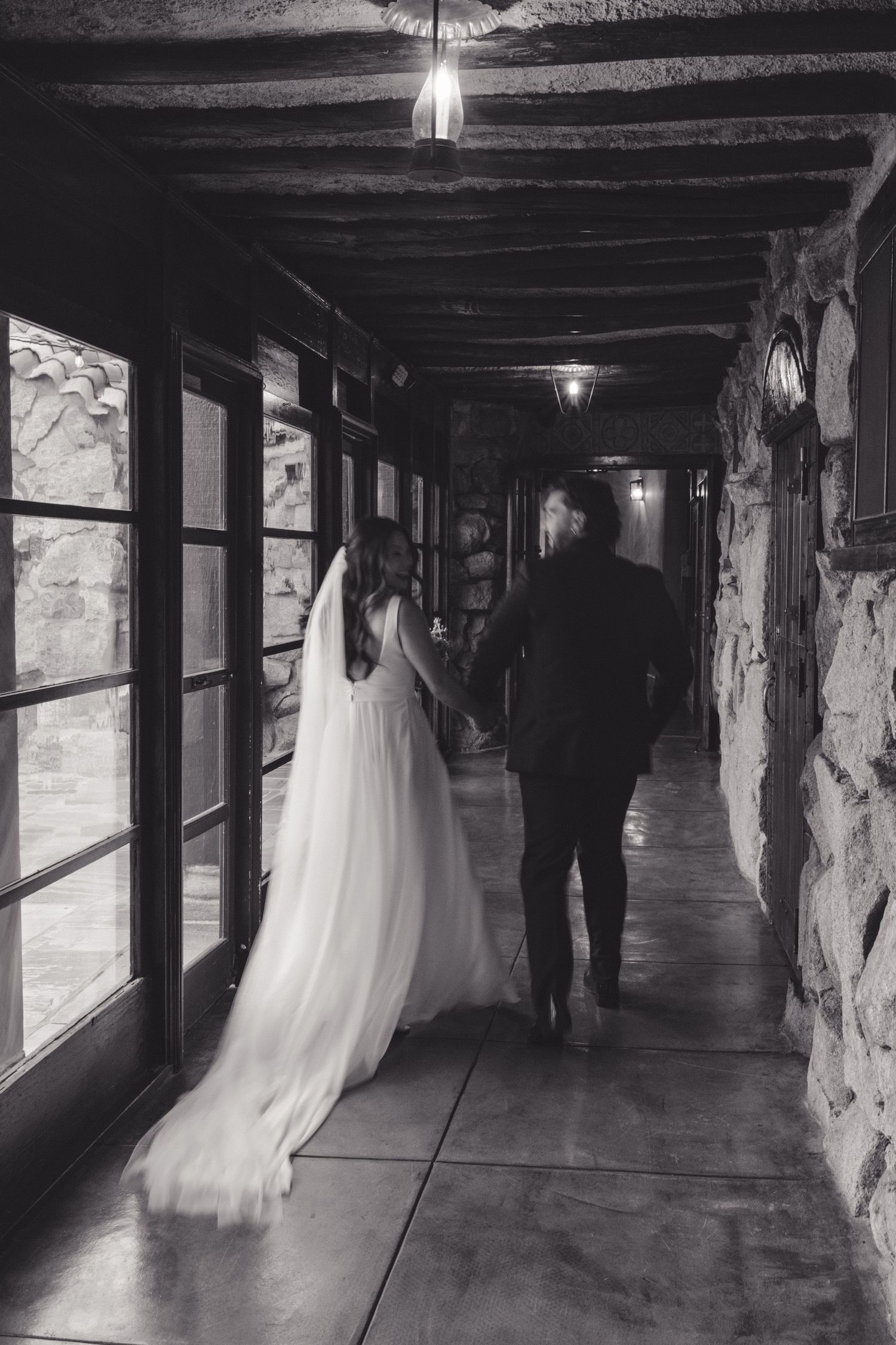 A couple walks down a dimly lit stone hallway with windows, the bride's flowing white dress trailing behind them.