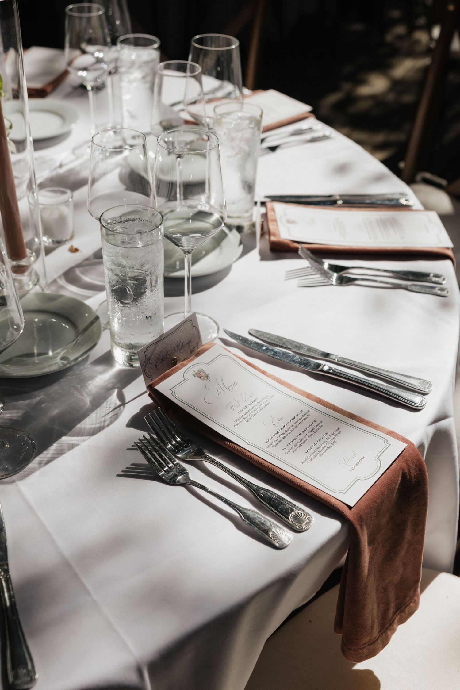 Elegant wedding reception table setting with white linen, silver cutlery, crystal glasses, and terracotta napkins with printed menus.