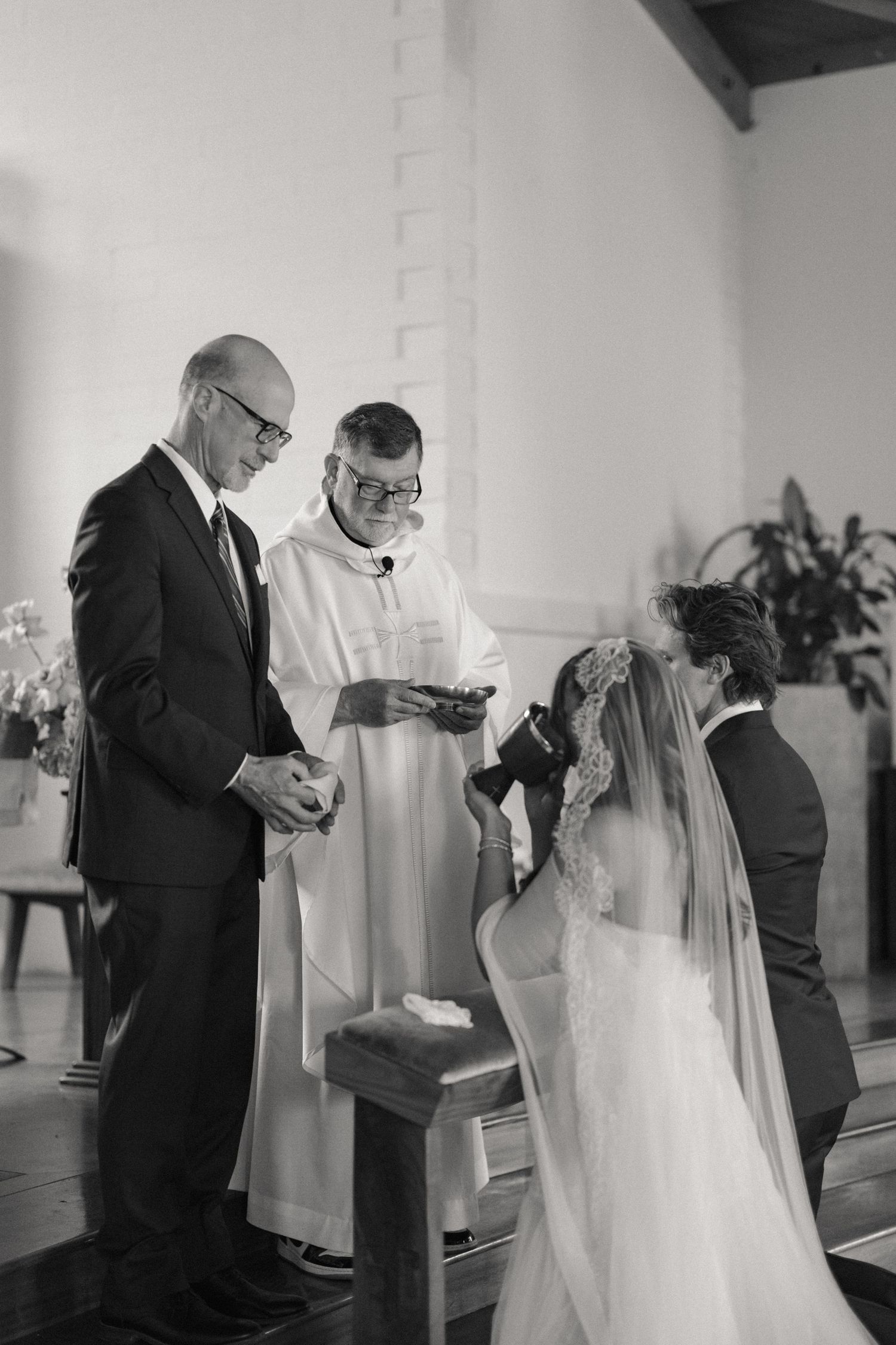 Black and white photo of bride receiving blessing from priest and father during church wedding ceremony.