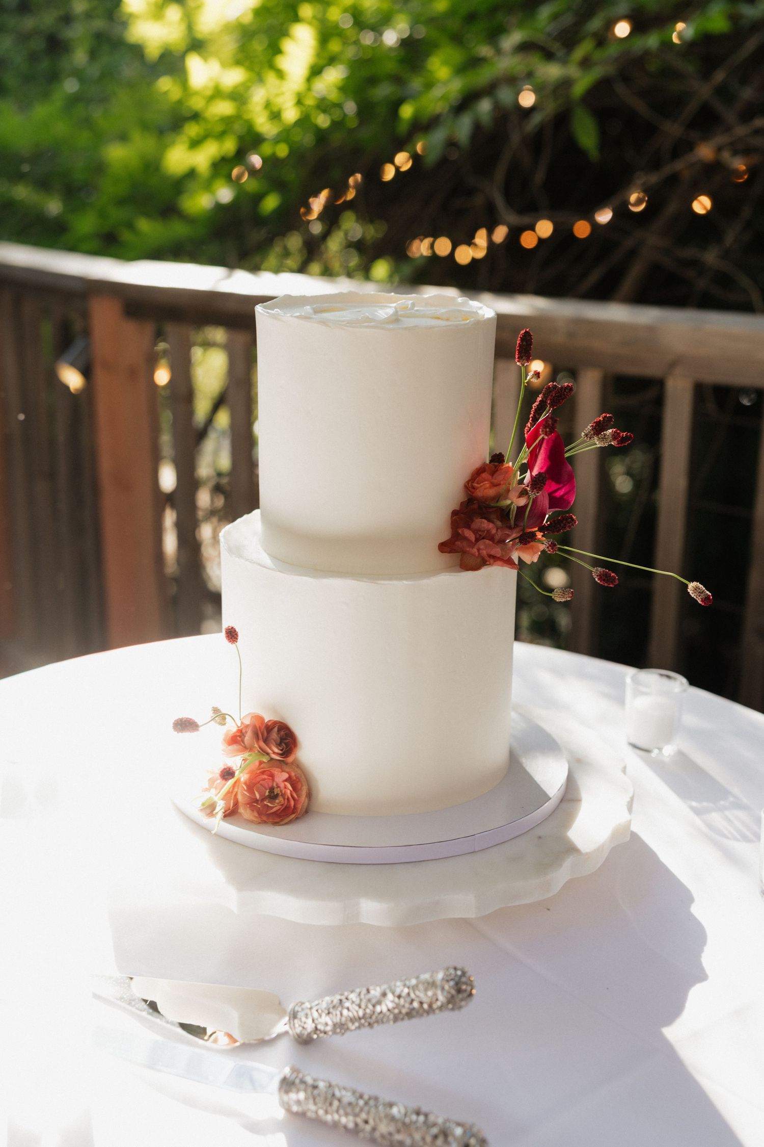 Elegant two-tier white wedding cake decorated with red and orange flowers on white tablecloth outdoors.