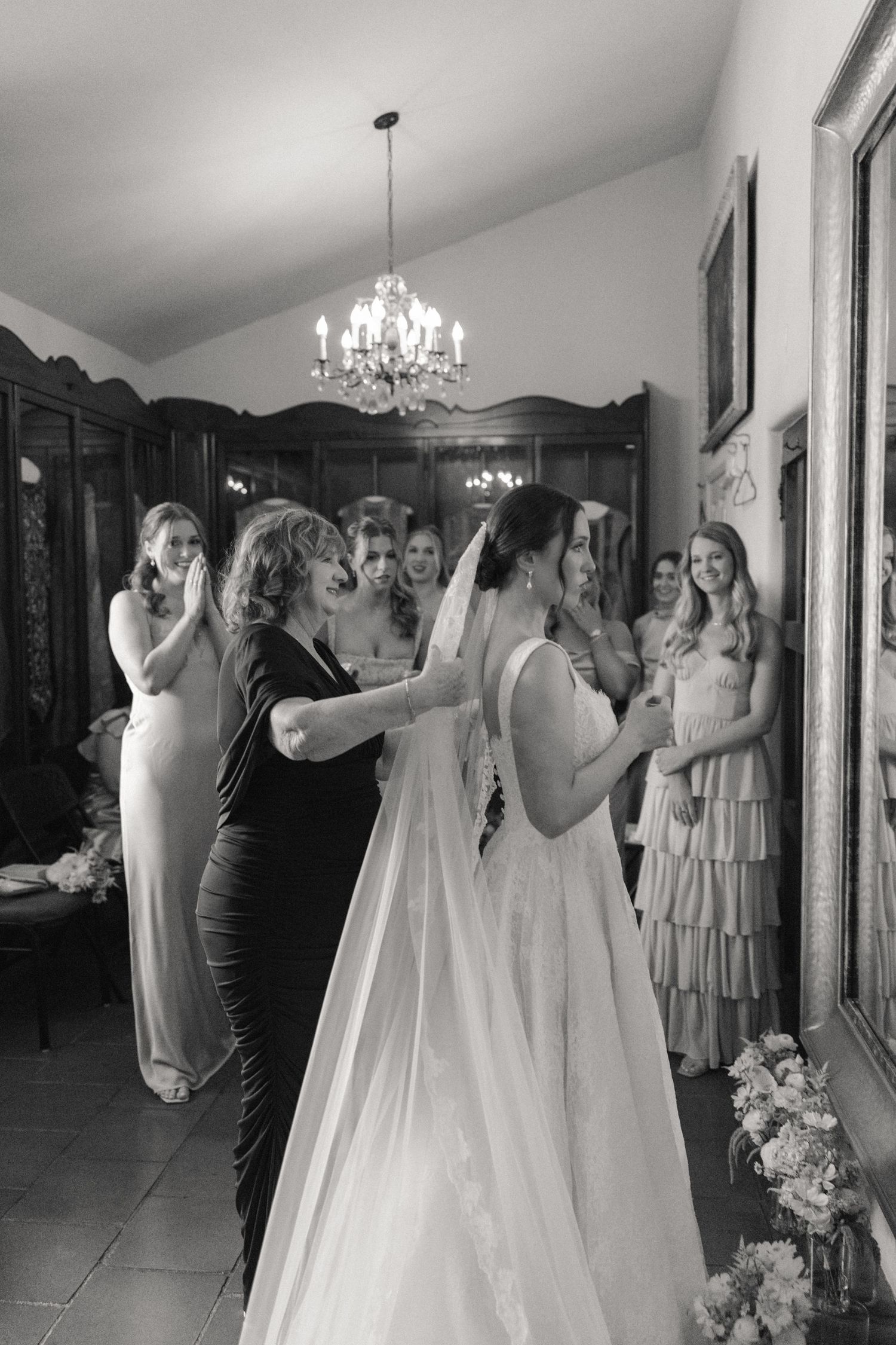 Black and white photo of bride in wedding gown and veil with bridesmaids and mother helping her prepare indoors.