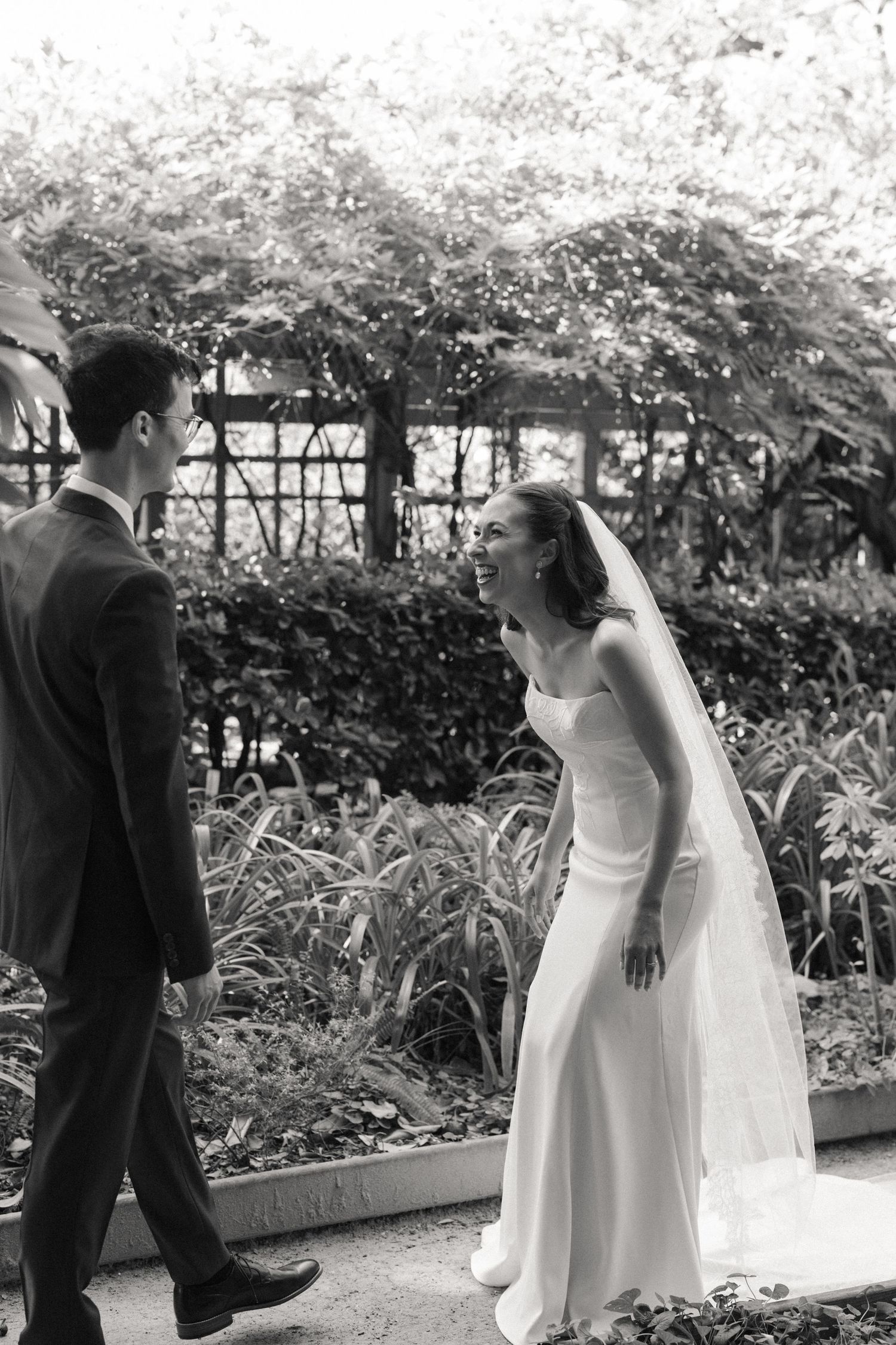 Black and white photo of a smiling bride in a white gown and veil facing a groom in a suit in a garden.
