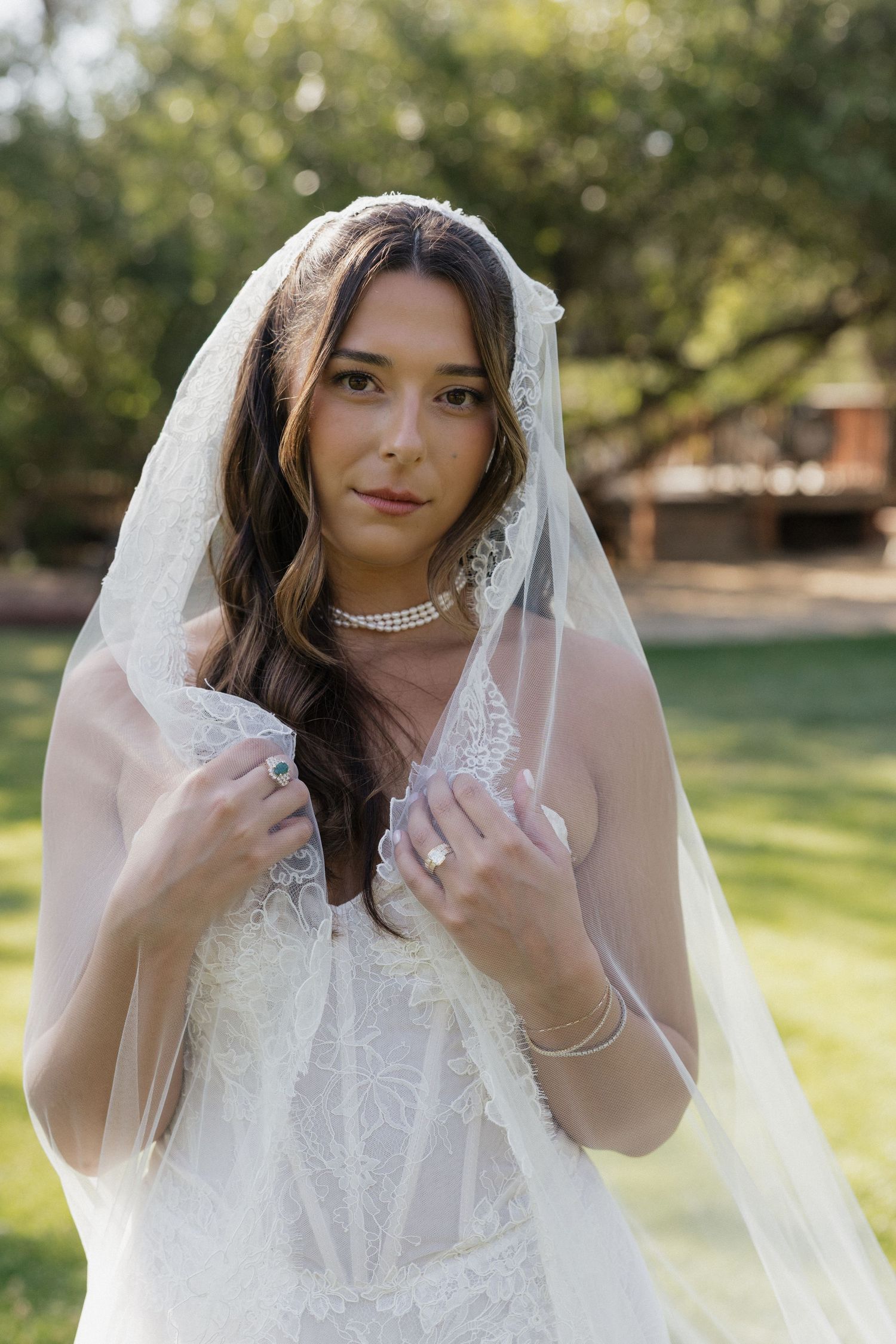 Elegant bride in lace wedding gown holding veil while posing outdoors in natural sunlight.
