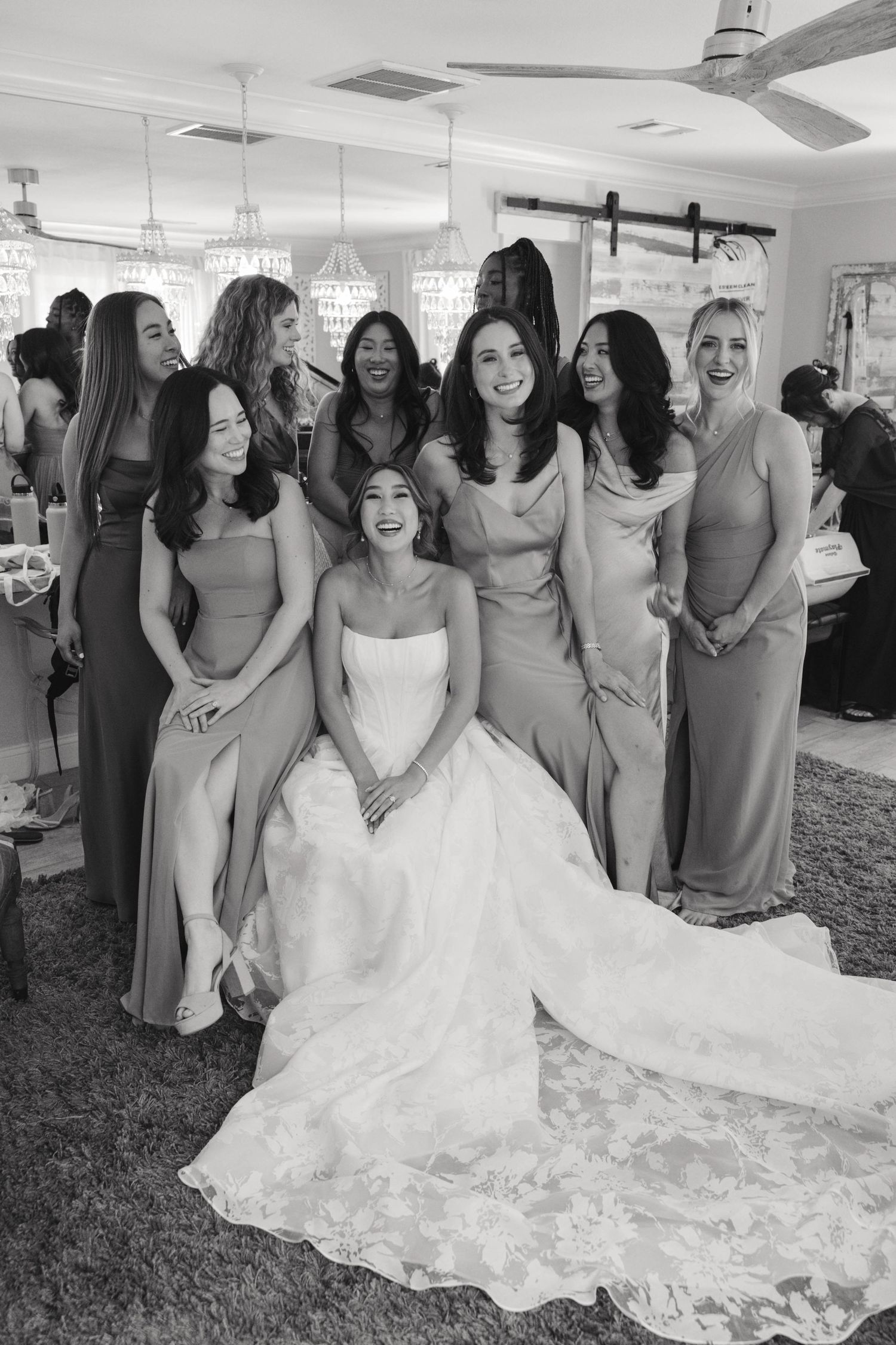 A bride in a flowing white gown shares joyful moments with bridesmaids wearing sage green dresses.