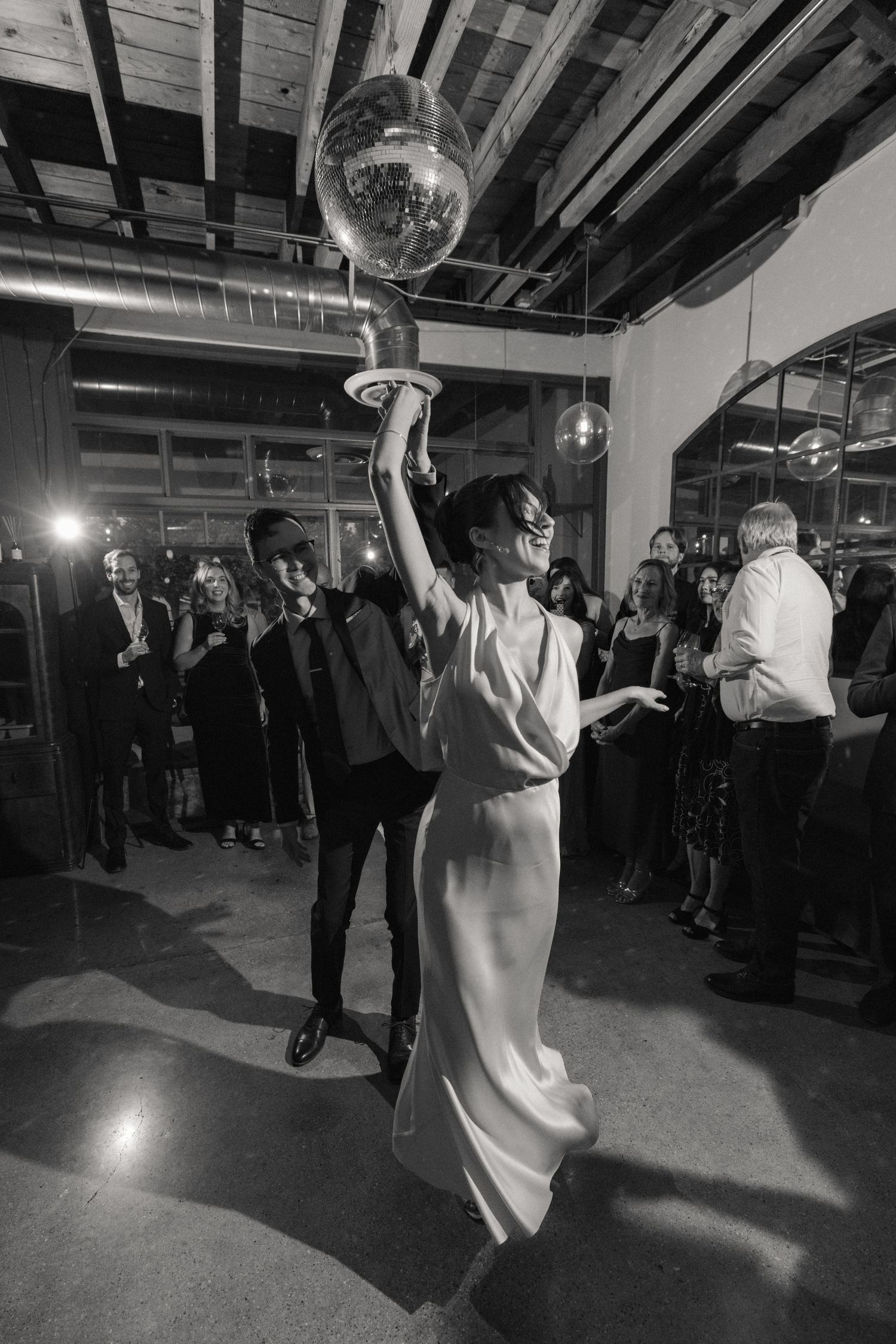 Bride in white gown holds disco ball aloft on dance floor at elegant wedding reception with guests watching.