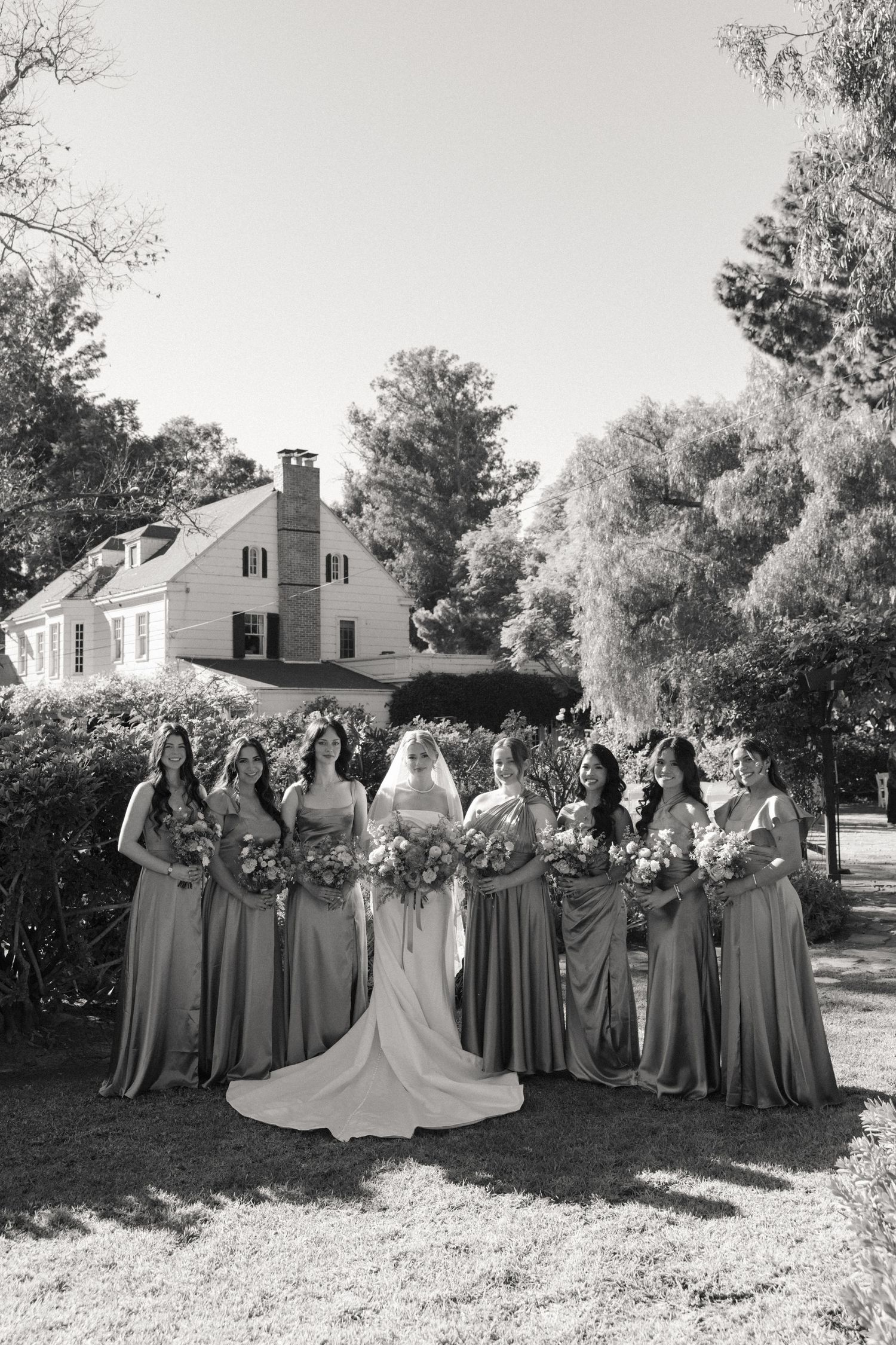 Wedding party in gray bridesmaid dresses poses outdoors in front of a white house on a sunny day.