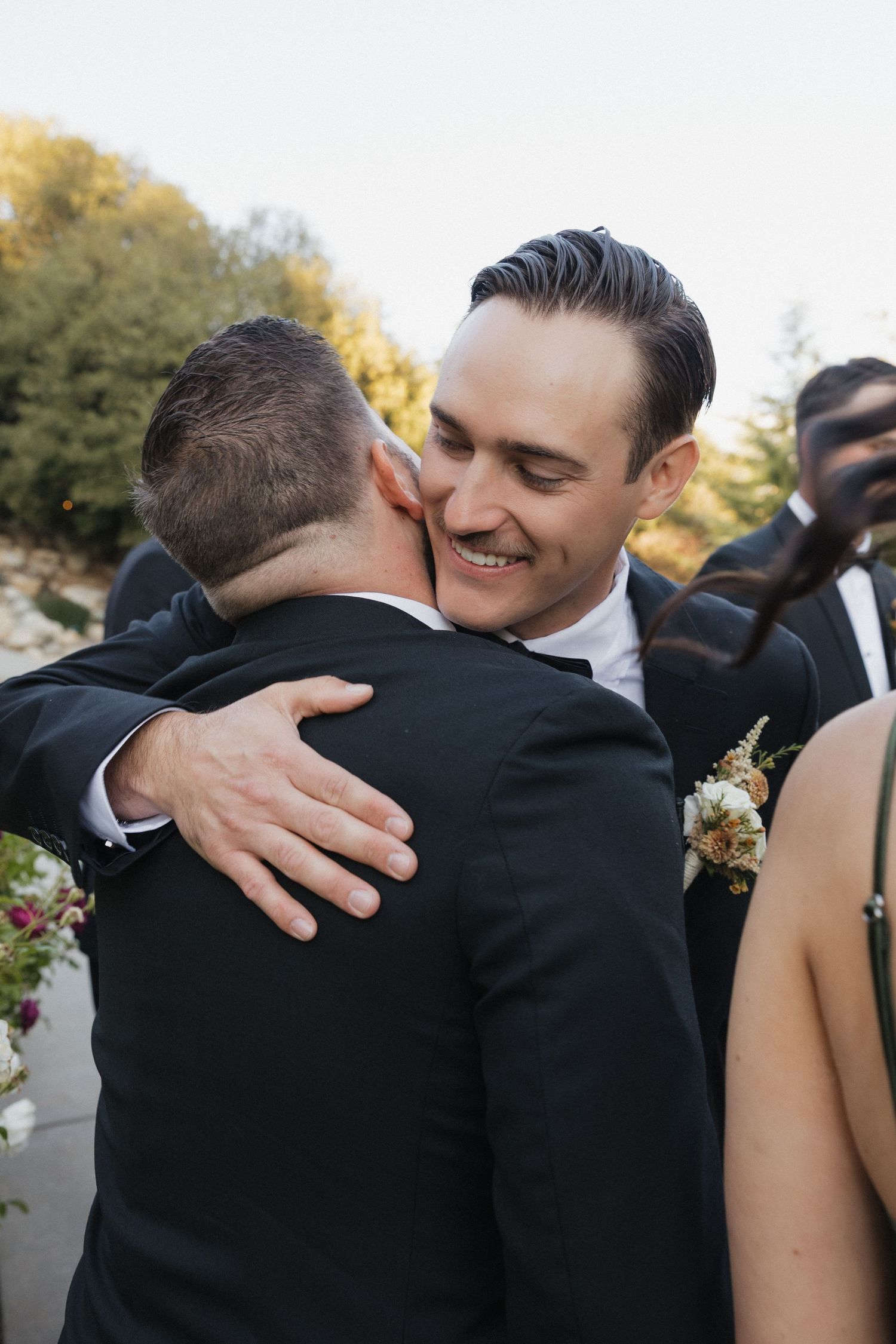 Groomsmen in black suits share emotional embraces during wedding party photos.