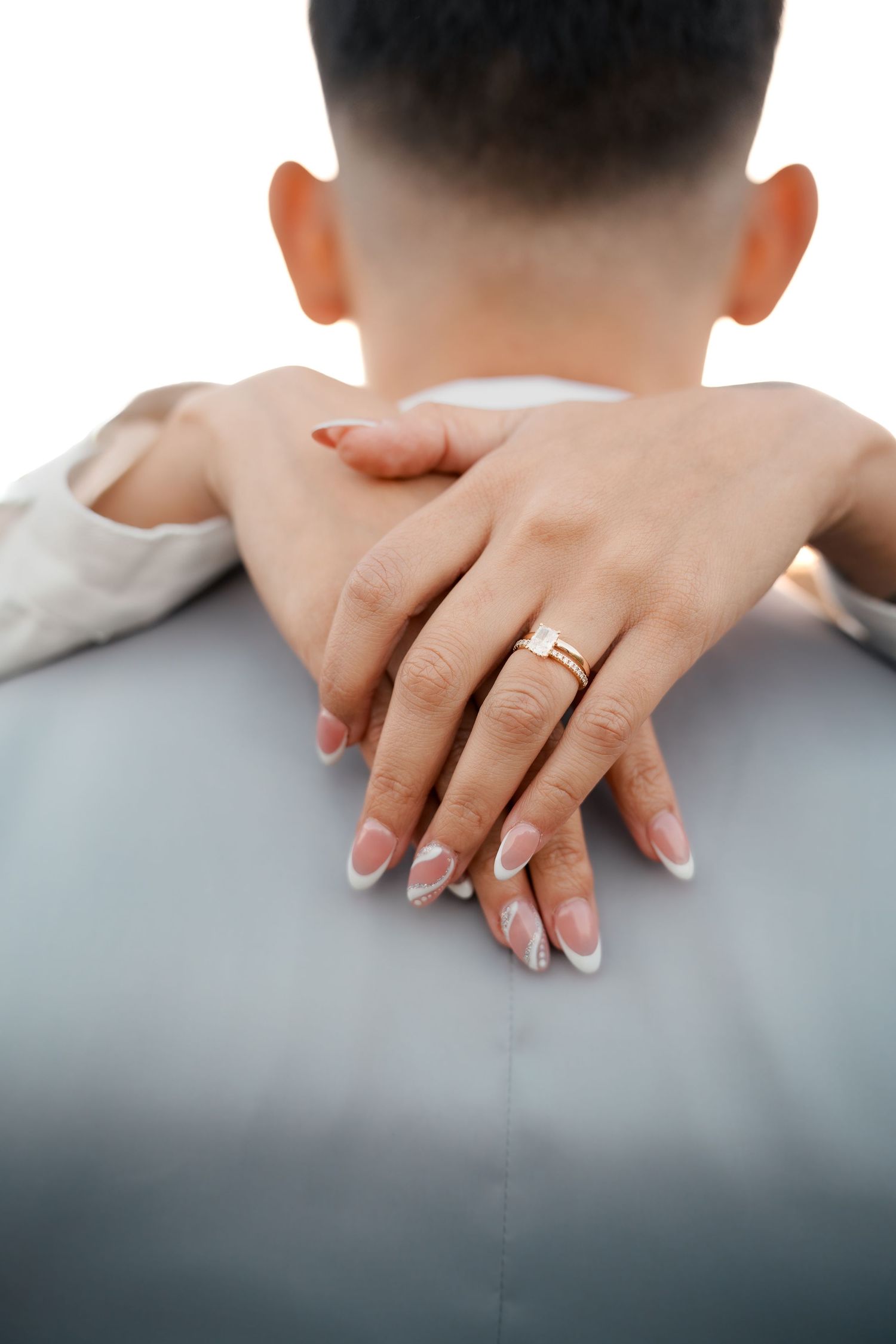 Close-up of hands with wedding ring embracing from behind, showing manicured nails.