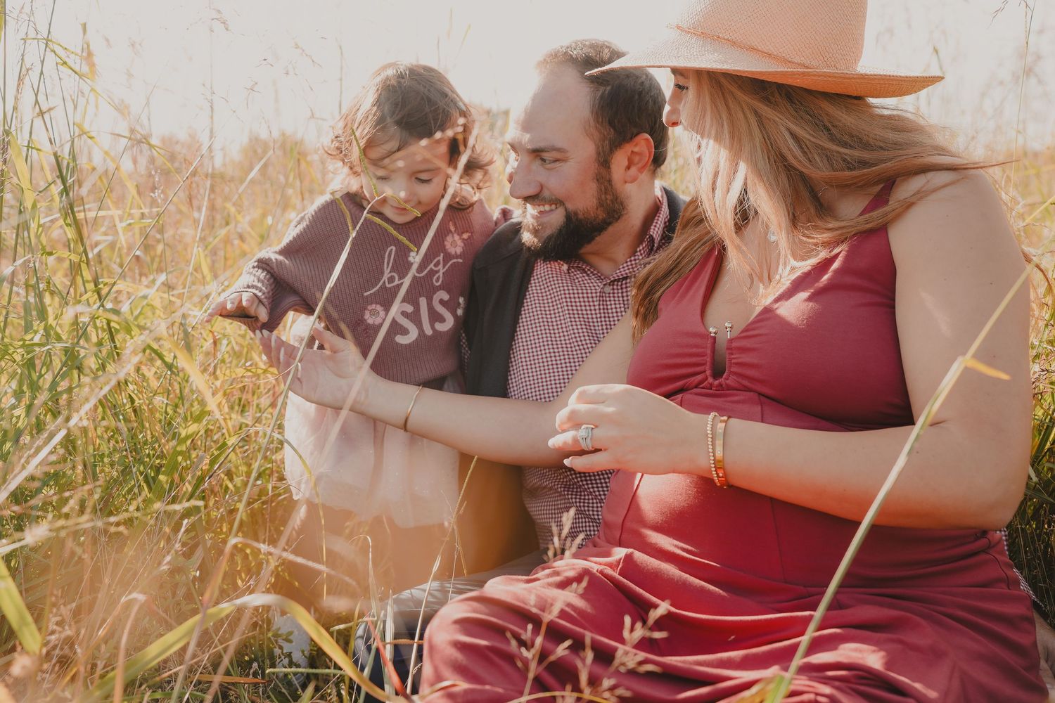 Three people sit together in a golden field of tall grass during sunset while sharing a casual outdoor moment.