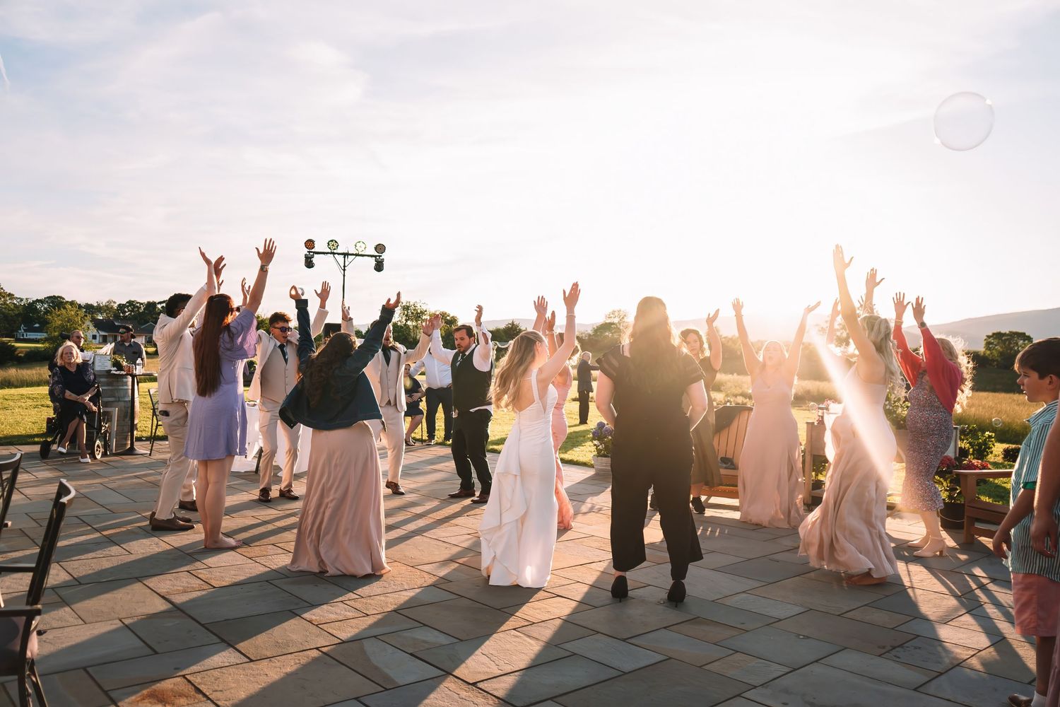 Wedding guests dance and celebrate with raised arms during an outdoor reception at sunset.