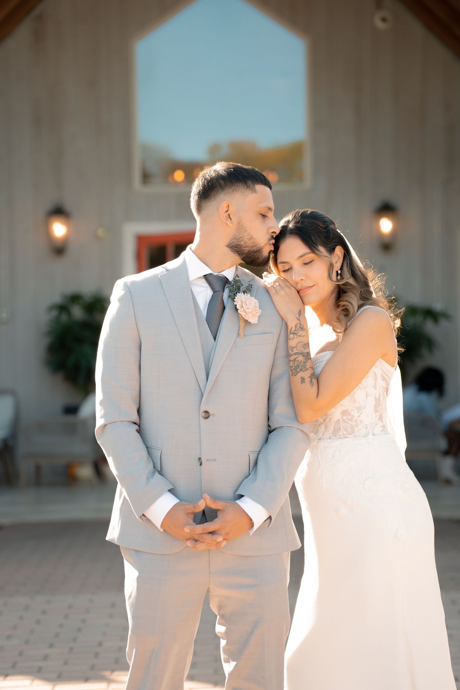 A romantic wedding photo of a couple in light gray and white attire standing together outside a chapel.