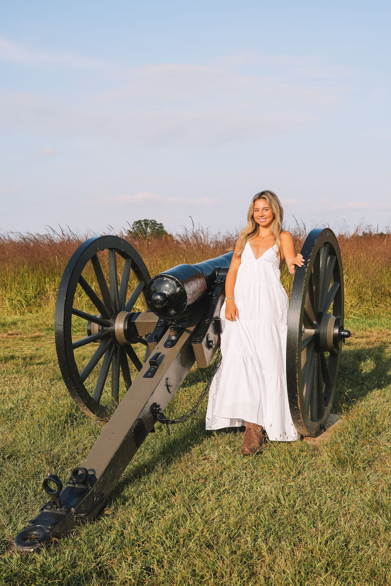 Graduation portrait session in the Manassas, Virginia in a white dress poses next to historic cannons on a grassy hilltop at sunset.