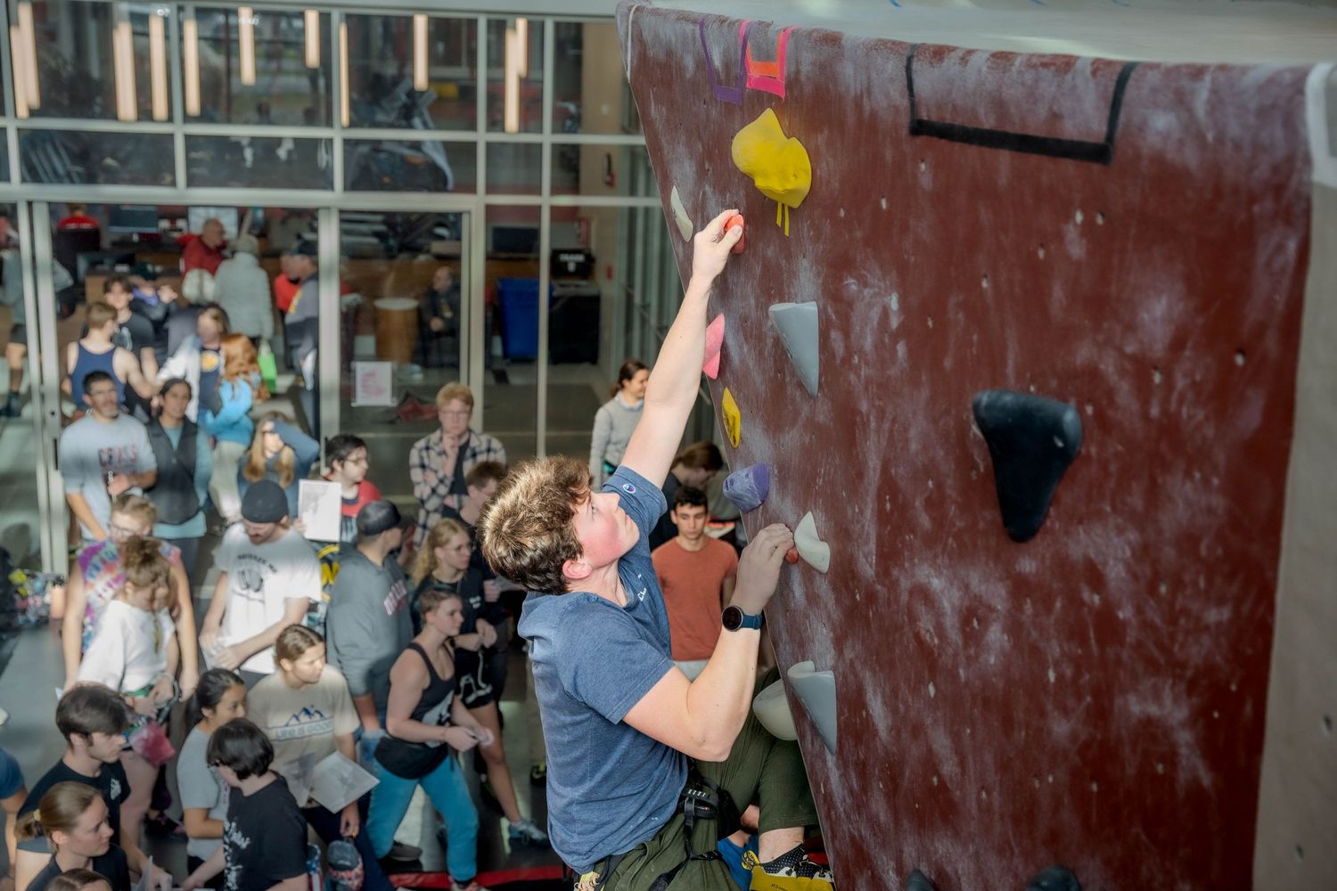 A rock climber stretches upward to reach a yellow hold on an indoor climbing wall while spectators watch from below.